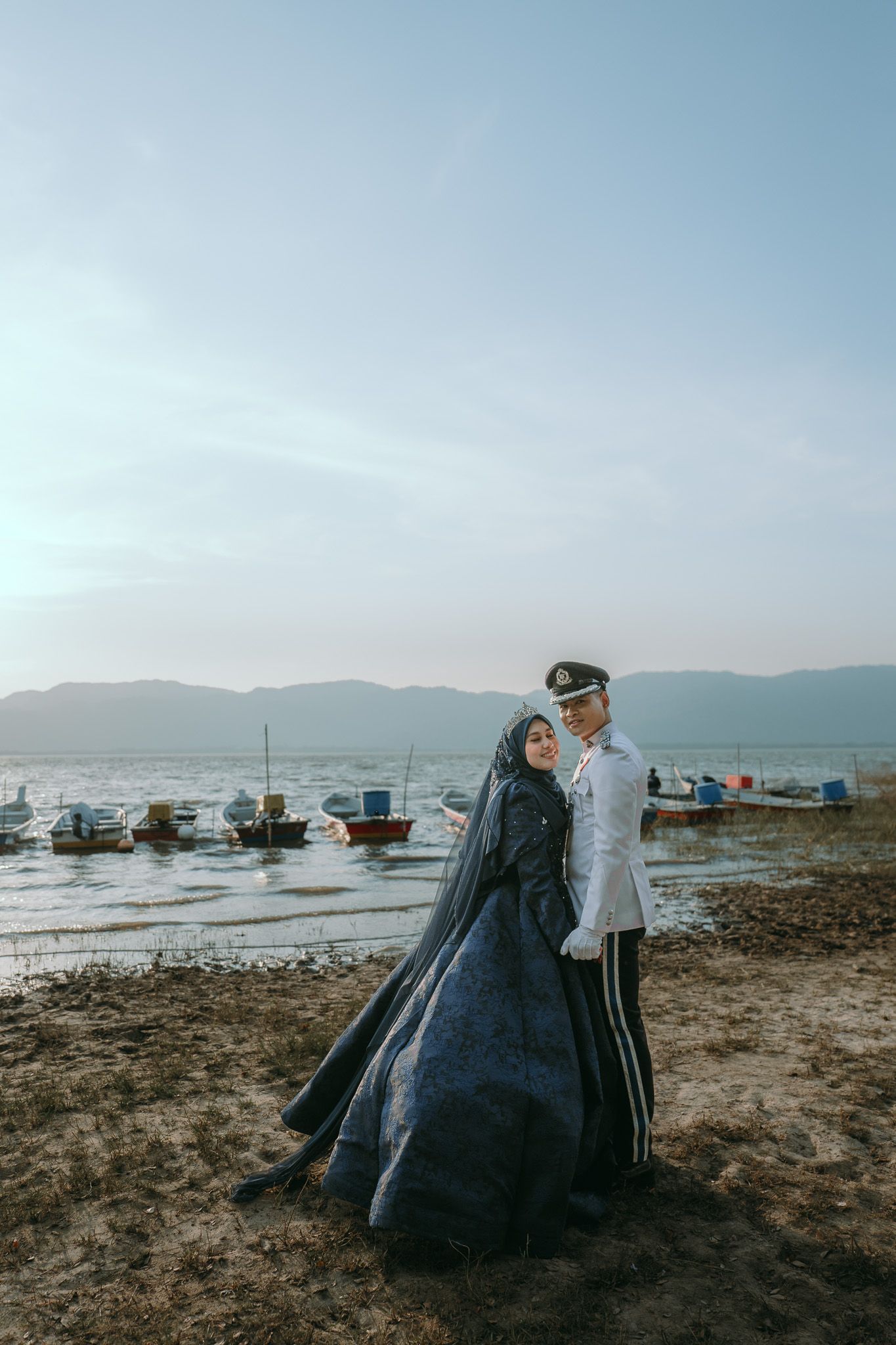 A couple in traditional dress stands on a sandy beach at sunset with fishing boats and mountains in the background.