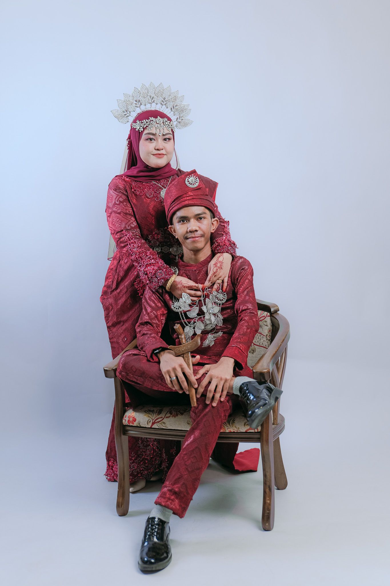 Two people wearing elegant burgundy outfits pose together on a wooden chair against a light gray background.