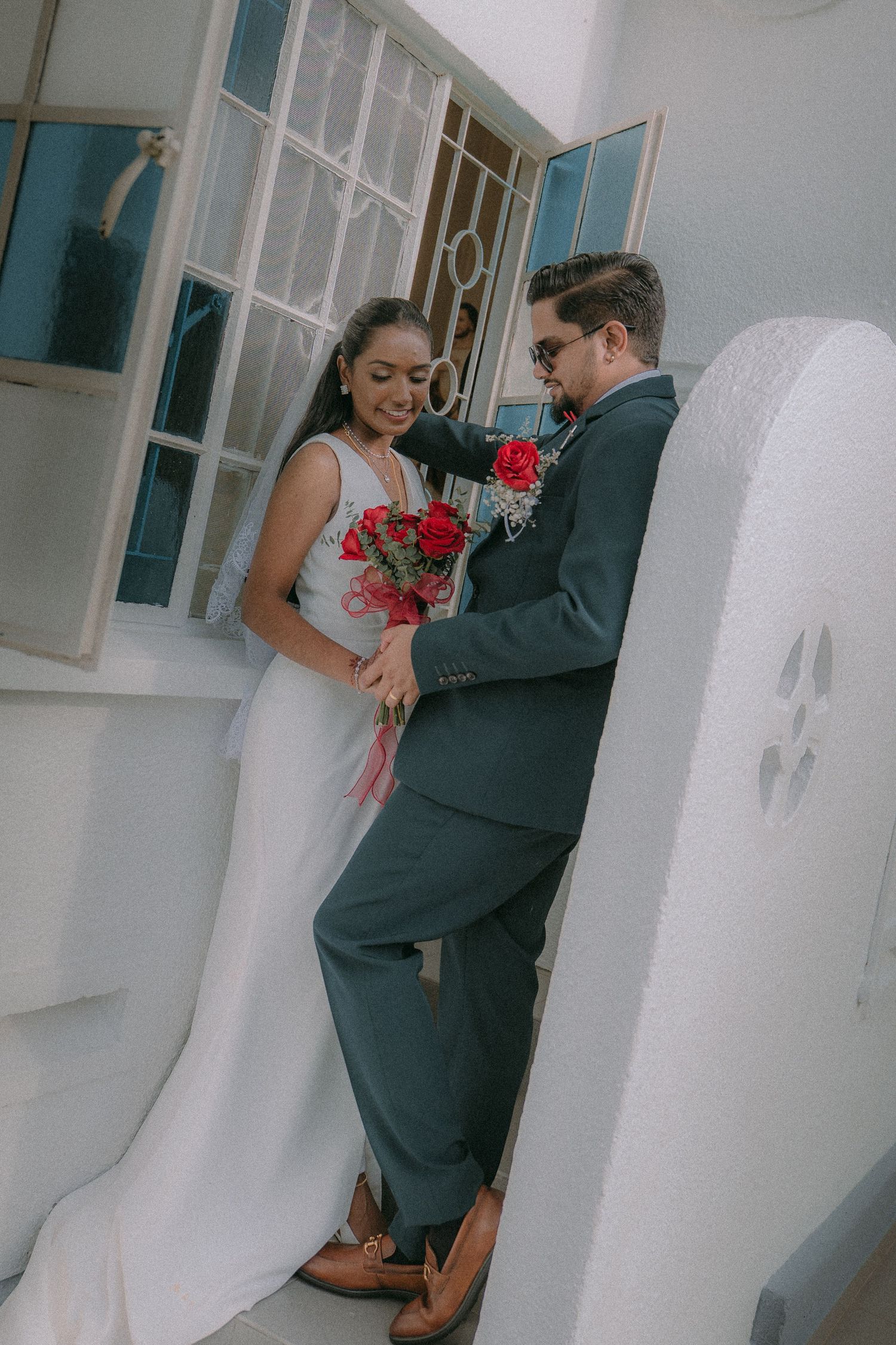 Romantic couple in bridal attire lean against white wall, holding red roses in intimate vintage-style setting.