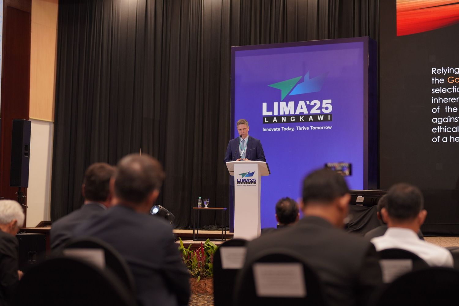 Speaker presenting at podium with LIMA25 branding displayed on screen in conference hall.