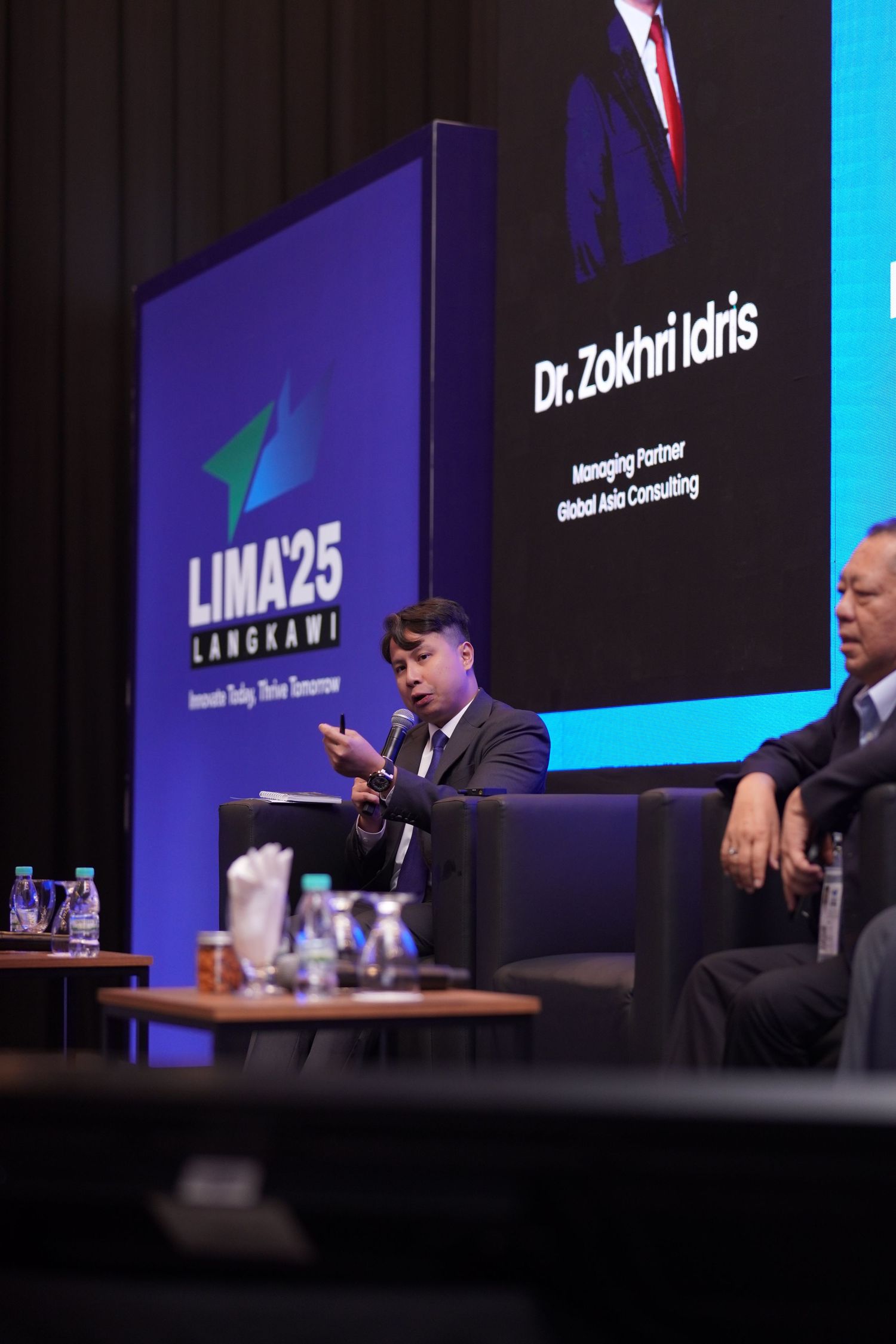 Conference speaker gestures on stage at LIMA25 leadership event with presentation screen visible.