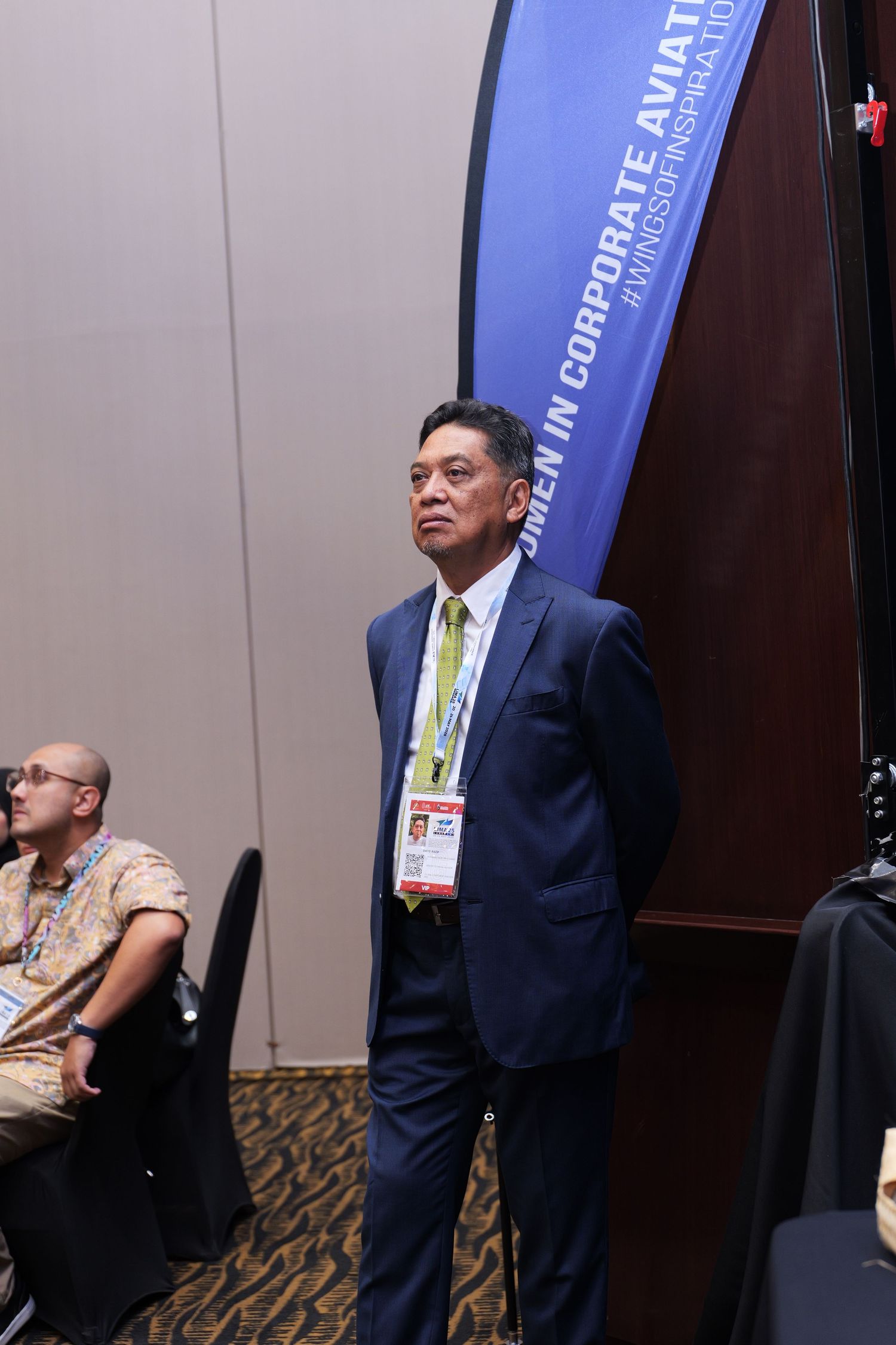 Business conference attendee wearing a navy suit and name badge standing in front of a blue banner display.