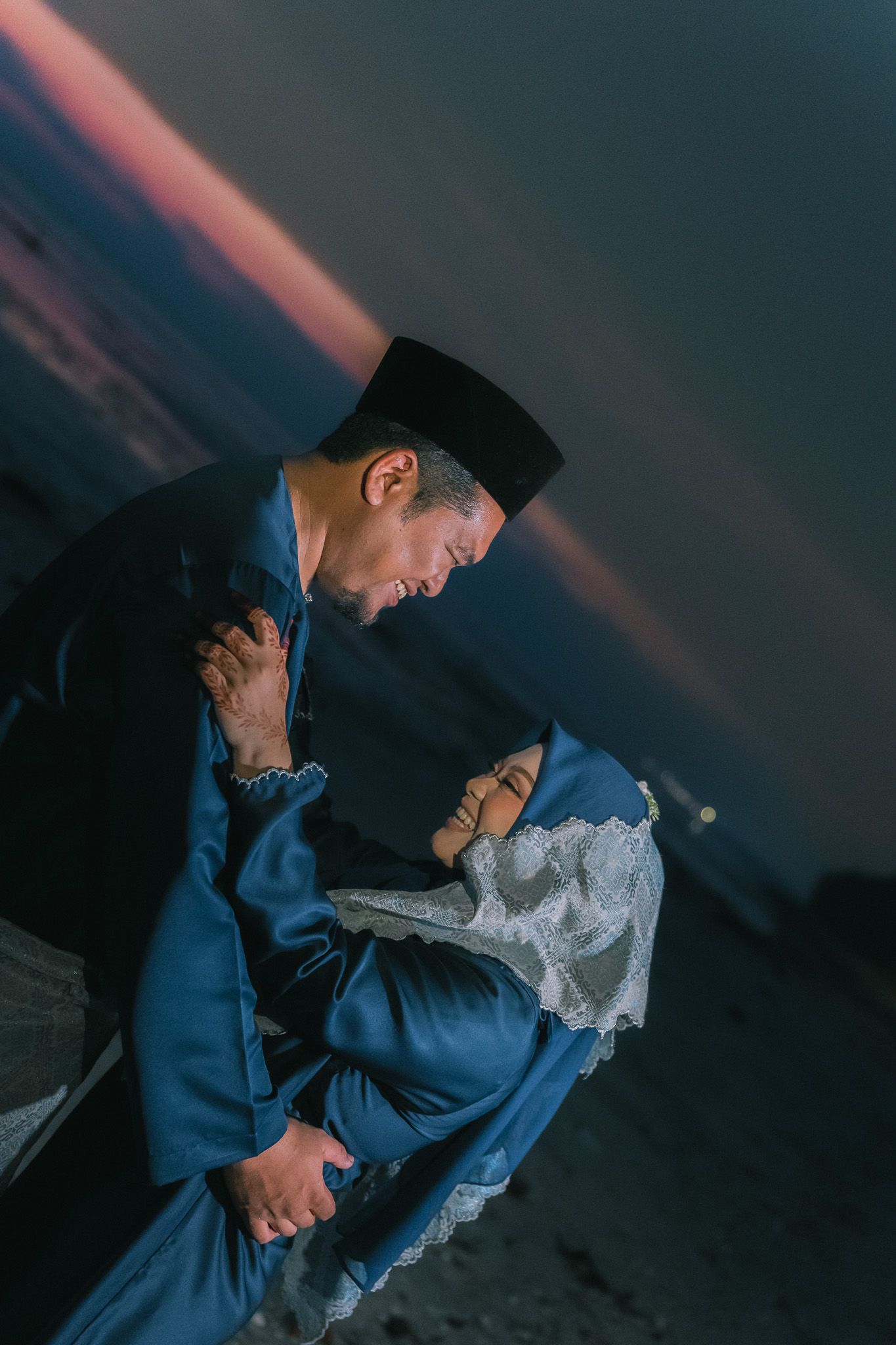 A person in traditional Muslim attire prays outdoors during sunset, bowing in reverence against a dark sky.