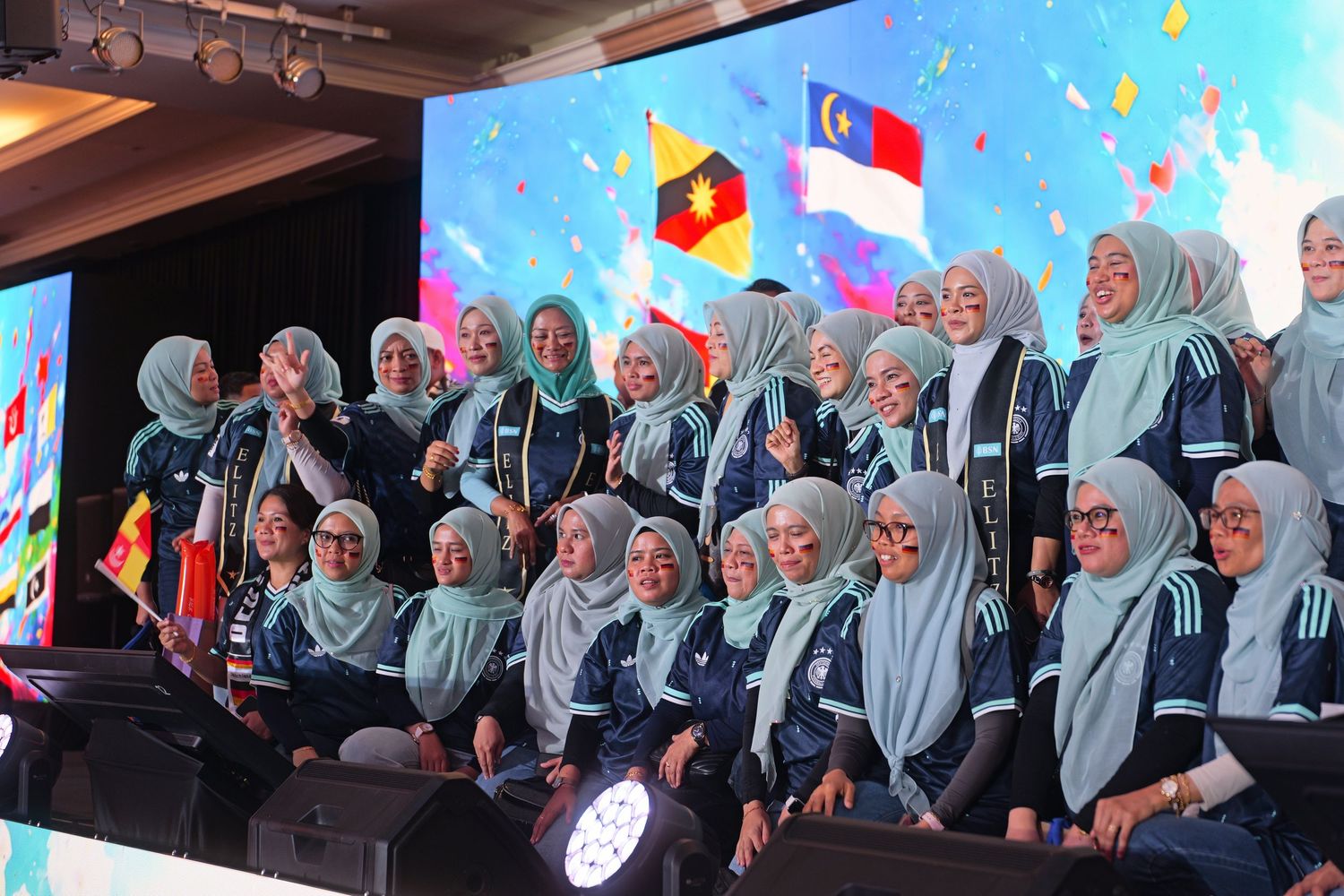 Group of women in matching hijabs and uniforms posing together at a Malaysian sporting event with flags in background.