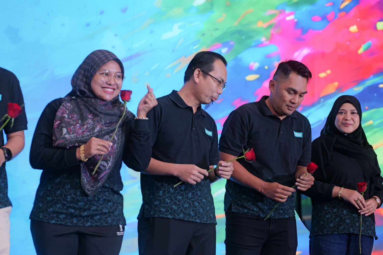 Group of people in matching black outfits smiling and holding roses at a corporate event on stage.