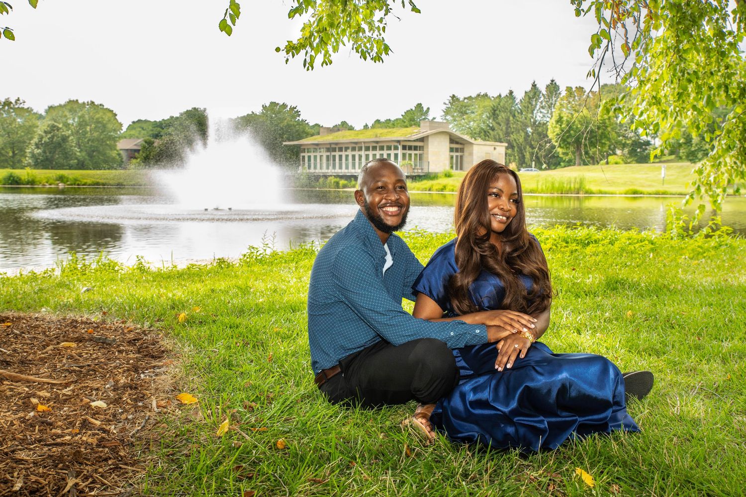 Couple sitting on green grass by a pond with a fountain, smiling, dressed in blue outfits outdoors.