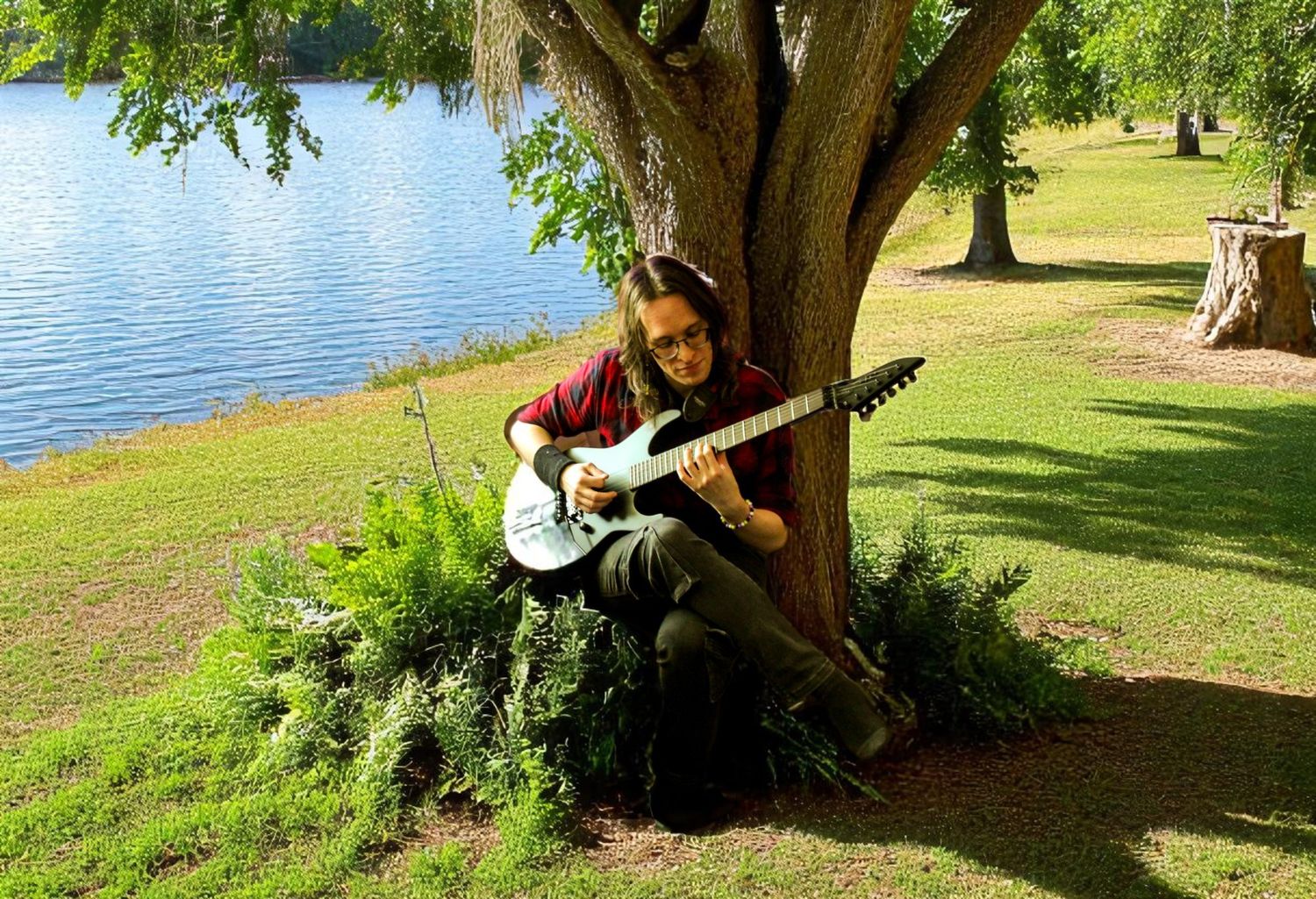 Person sitting under a large tree by a lake playing guitar on a sunny day.