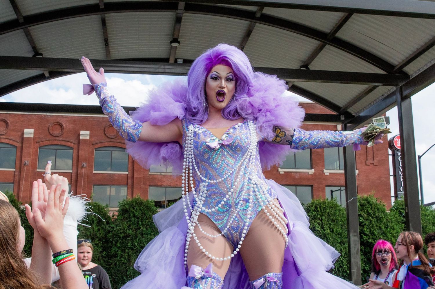 Performer in lavender costume and feathers dances energetically during outdoor stage performance.