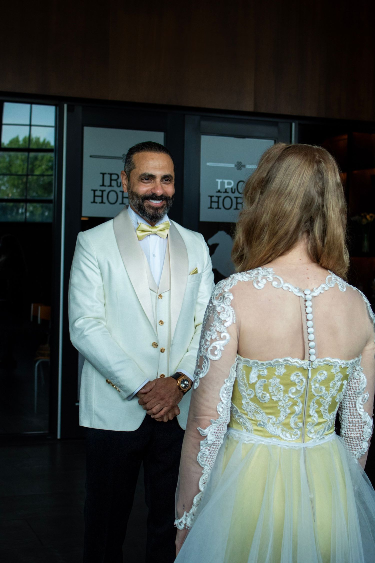 A wedding couple stands together, with the bride wearing a yellow and white lace dress and the groom in a white tuxedo.