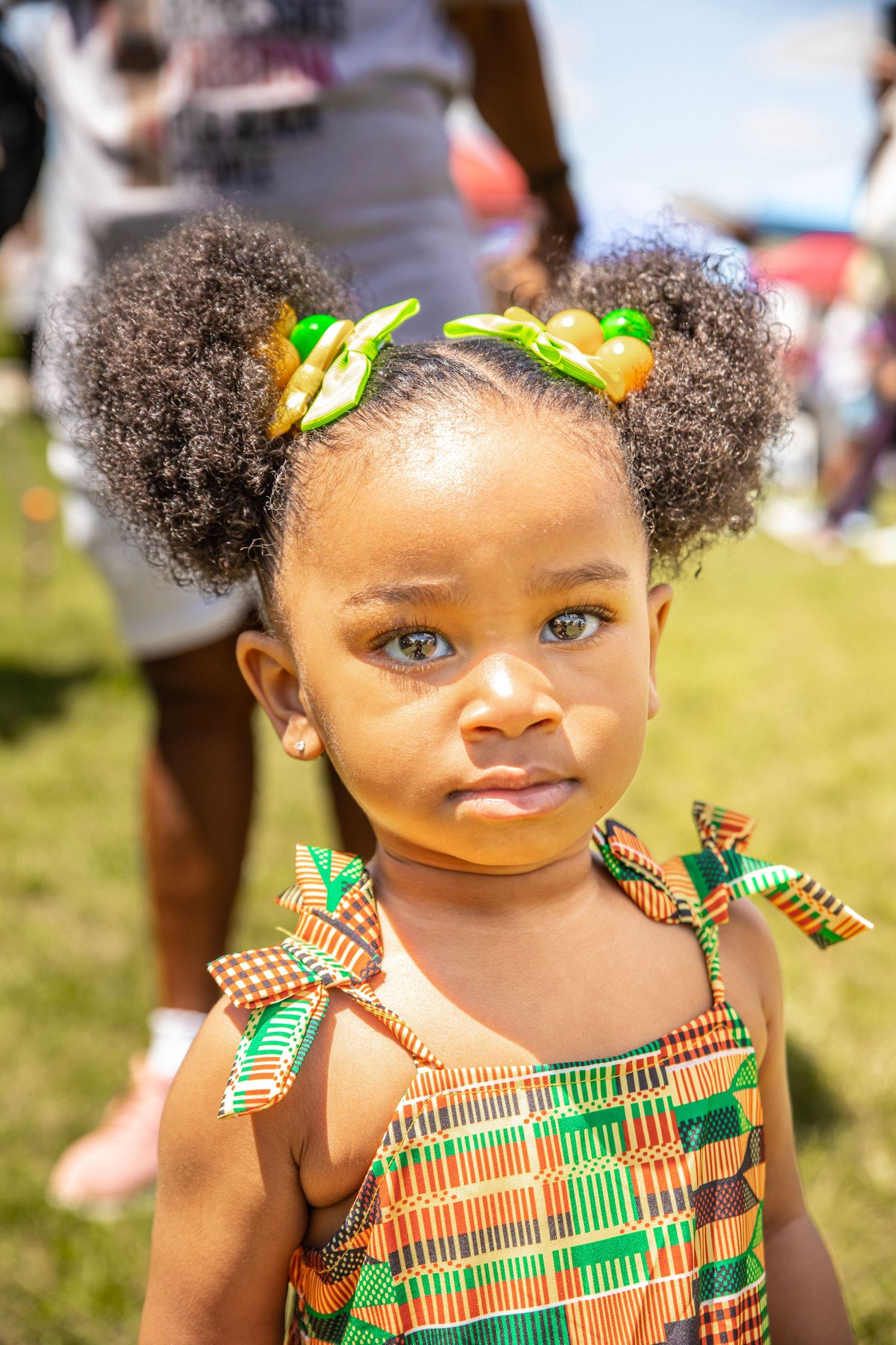 Young girl in colorful Kente print dress with afro puffs and green hair accessories at outdoor event.