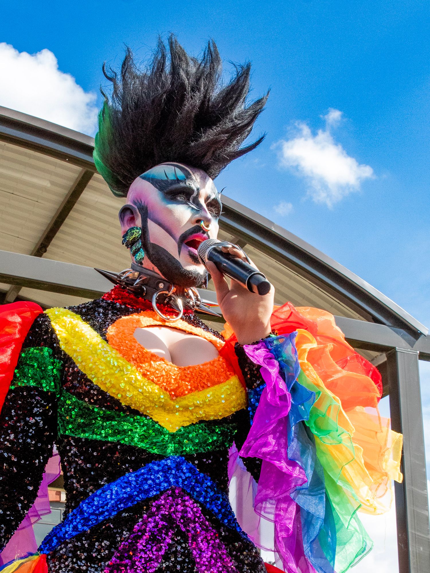 Performer in rainbow sequined dress and spiked mohawk hairstyle sings against blue sky background at outdoor event.