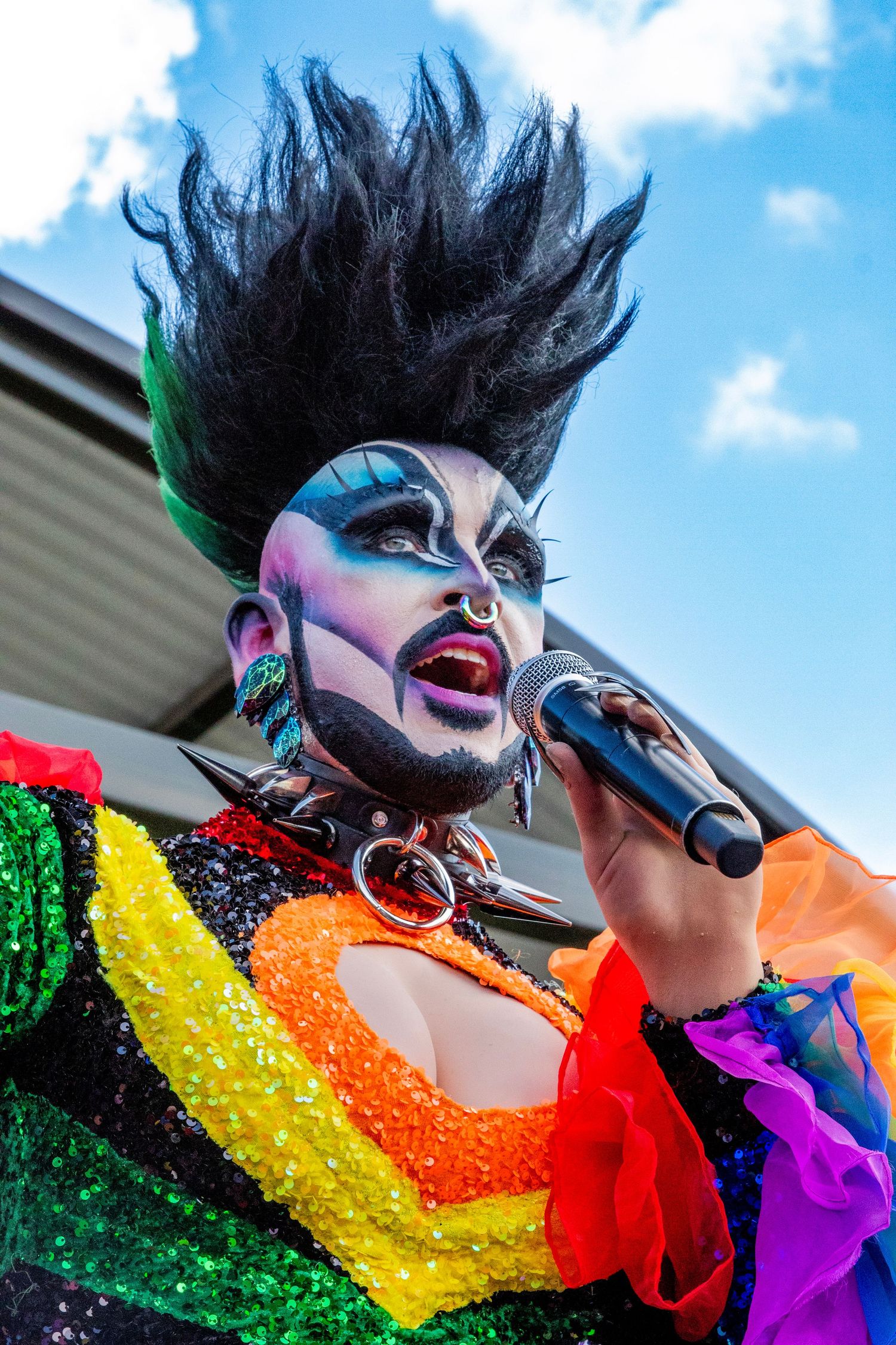 Drag performer in dramatic makeup and spiked black hair wearing a rainbow sequined outfit performs with a microphone outdoors.