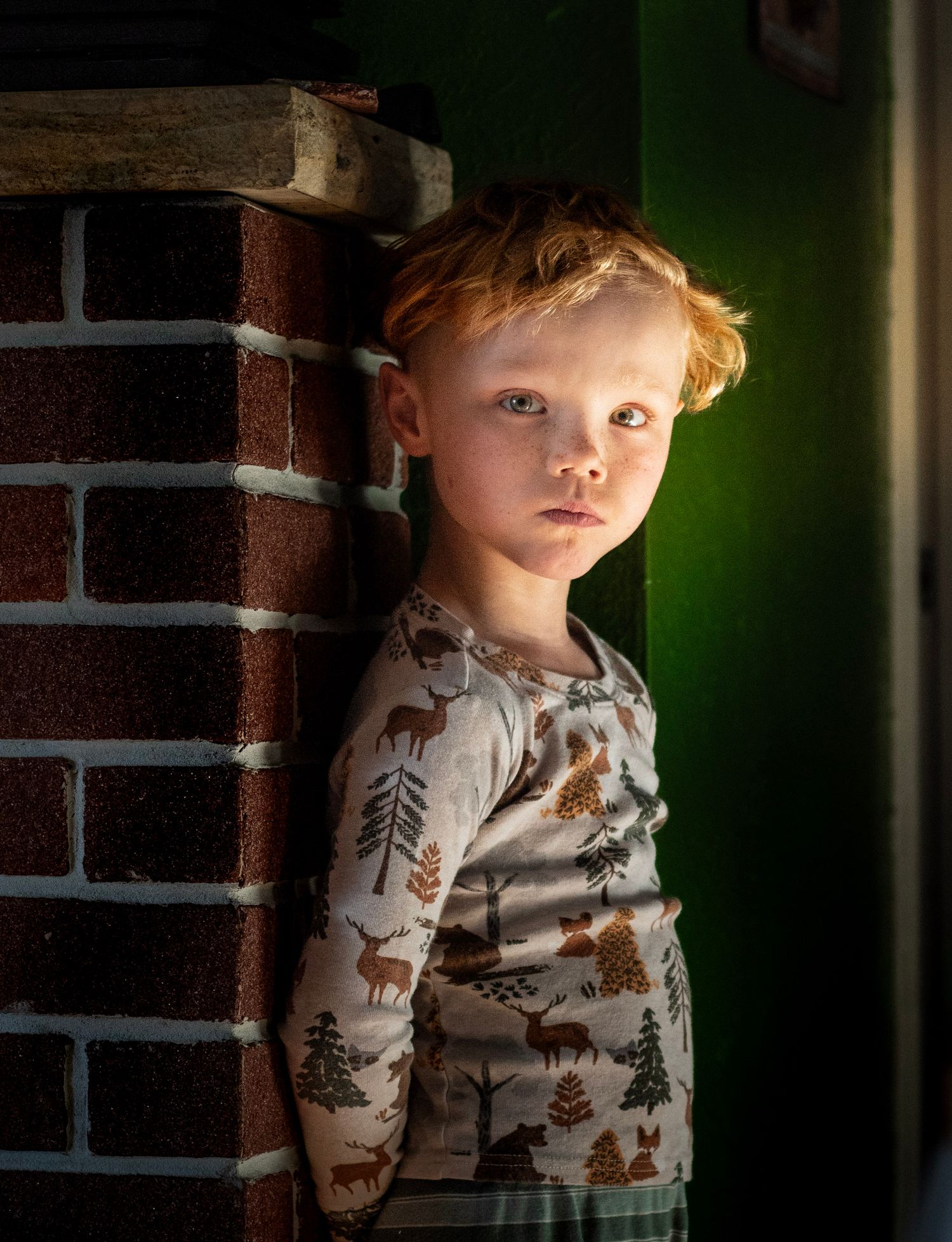 Red-haired child in forest-print pajamas leaning against a brick wall with dramatic green and golden lighting.