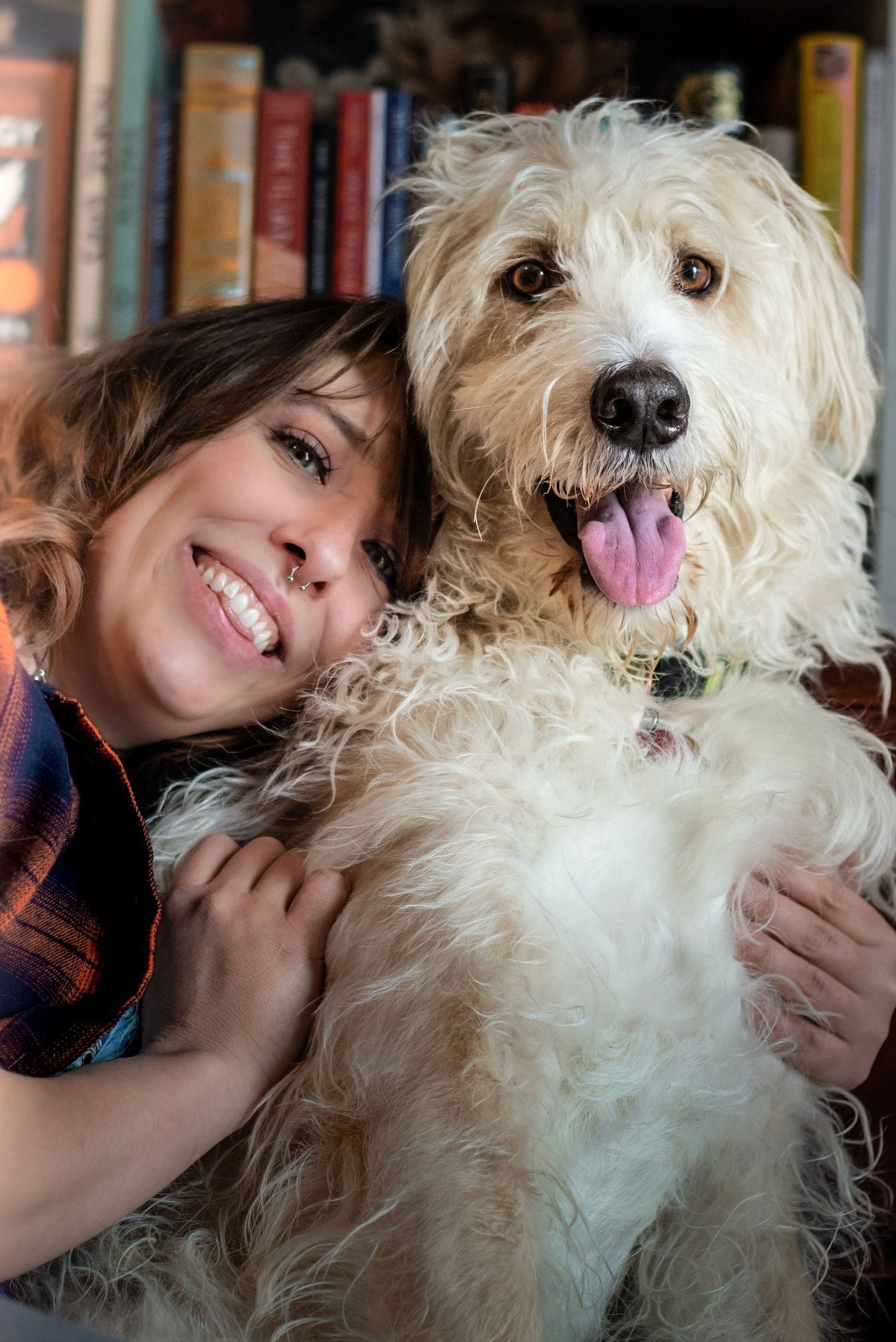 Smiling woman hugging a fluffy white Goldendoodle dog in front of a bookshelf.