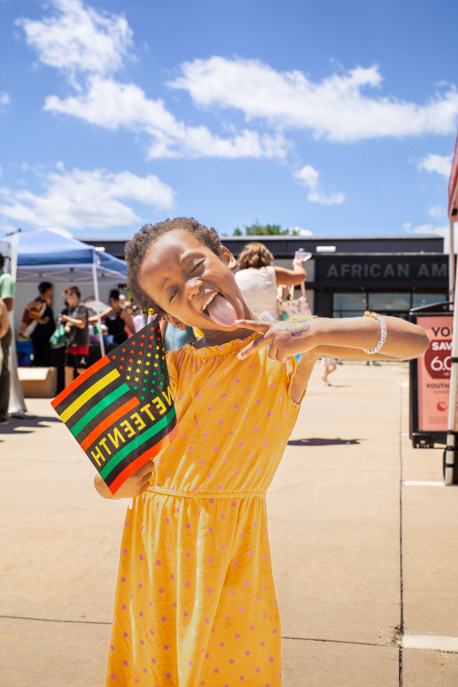 Young girl in yellow dress joyfully holds a Juneteenth flag at an outdoor African American cultural celebration.