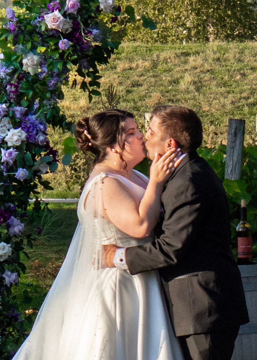Bride and groom share their first kiss at outdoor wedding ceremony beneath a purple floral arch.