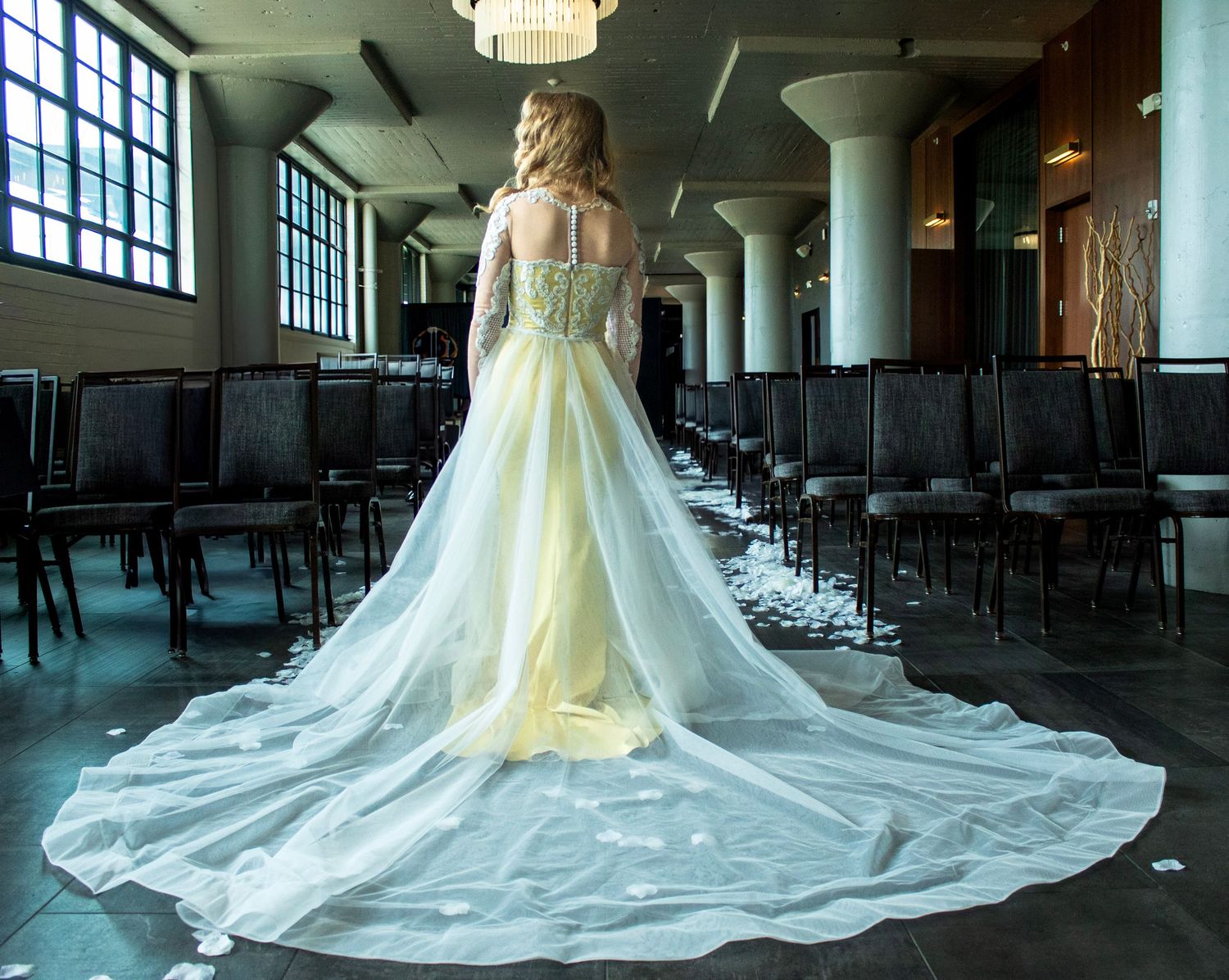 An elegant white wedding gown with cathedral-length train displayed in a spacious venue with large windows.