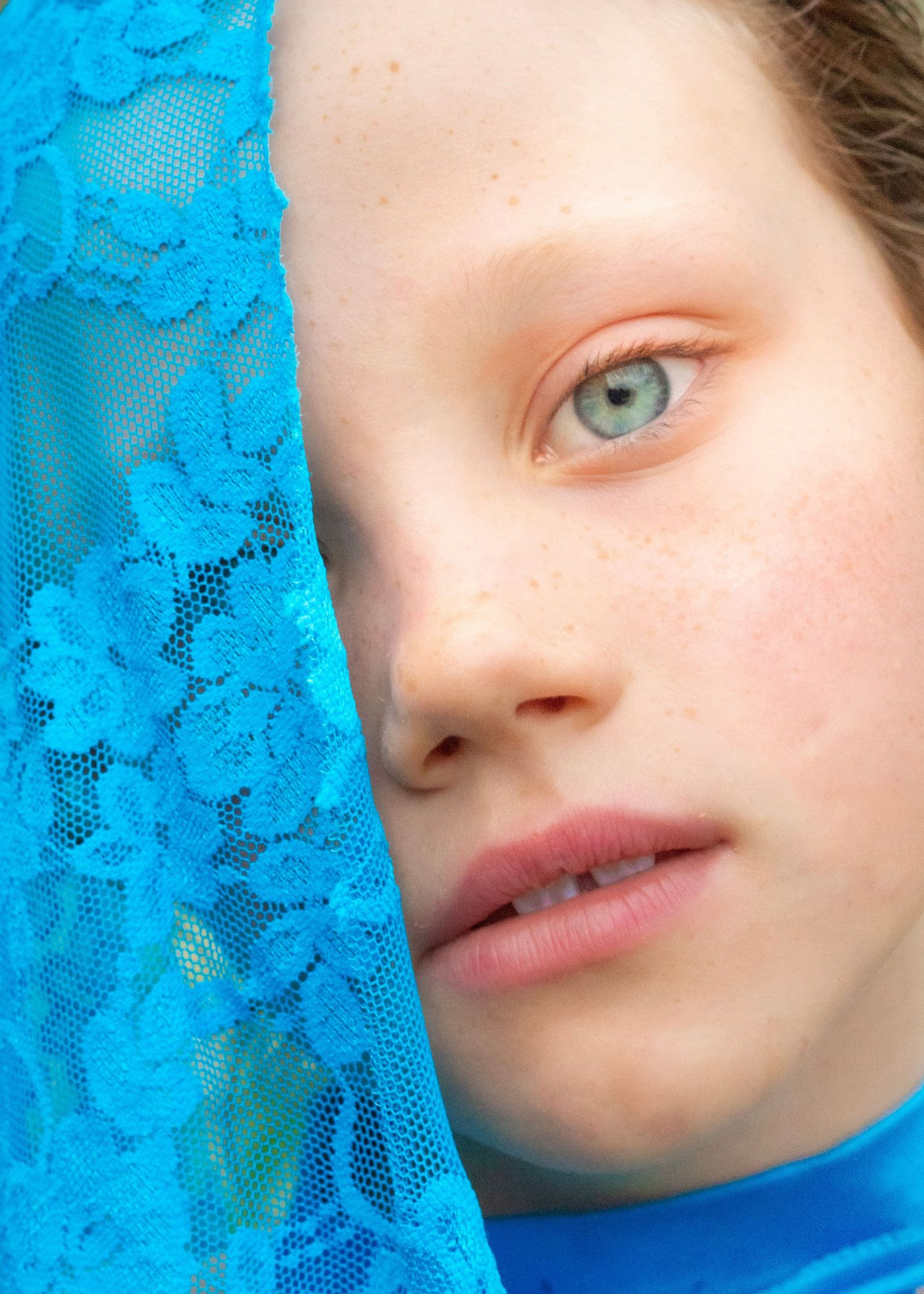 Close-up portrait of a freckled child with blue-green eyes, half-face obscured by blue floral lace fabric.