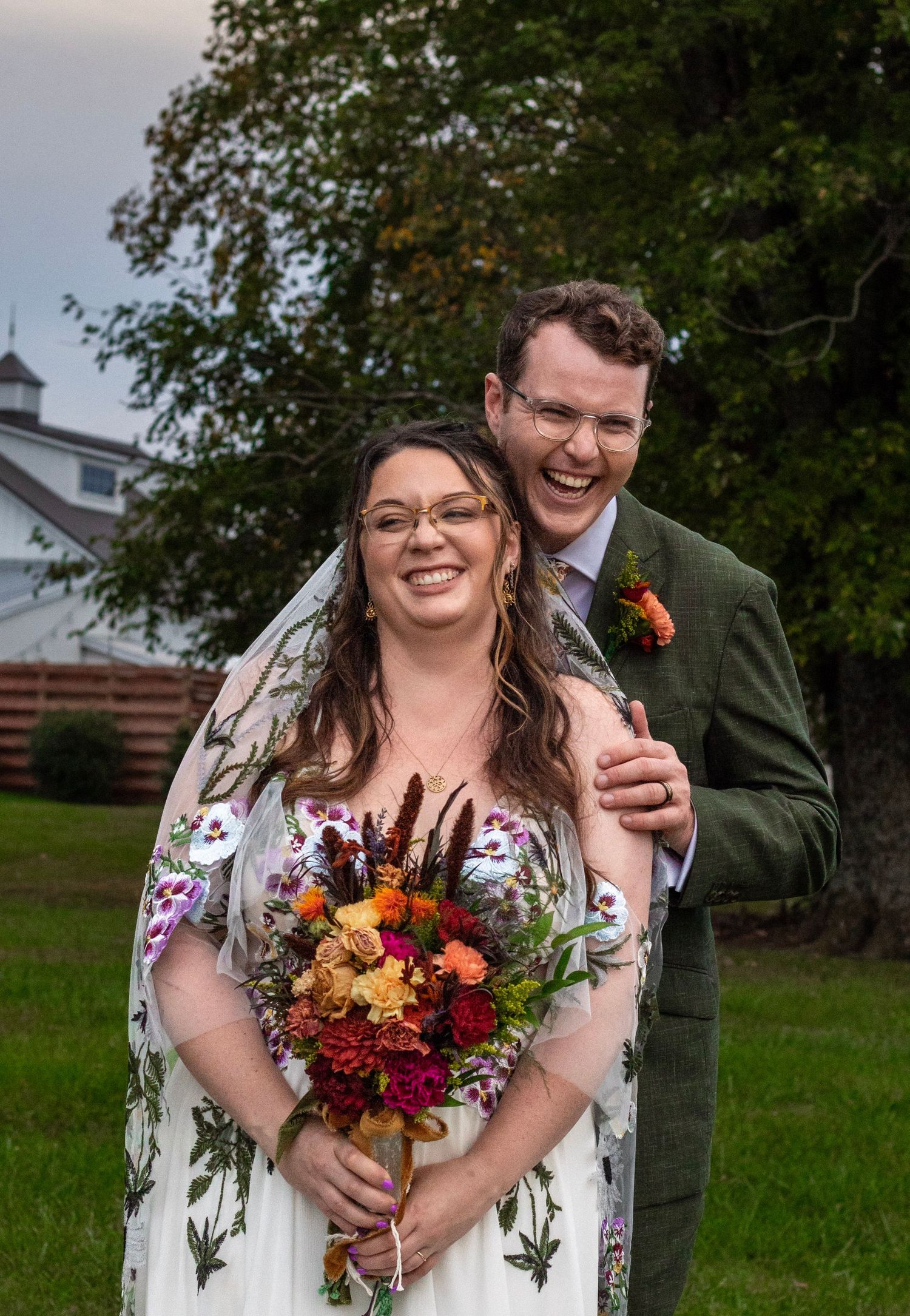 Smiling bride in floral wedding dress holding colorful bouquet with groom in green suit outdoors.