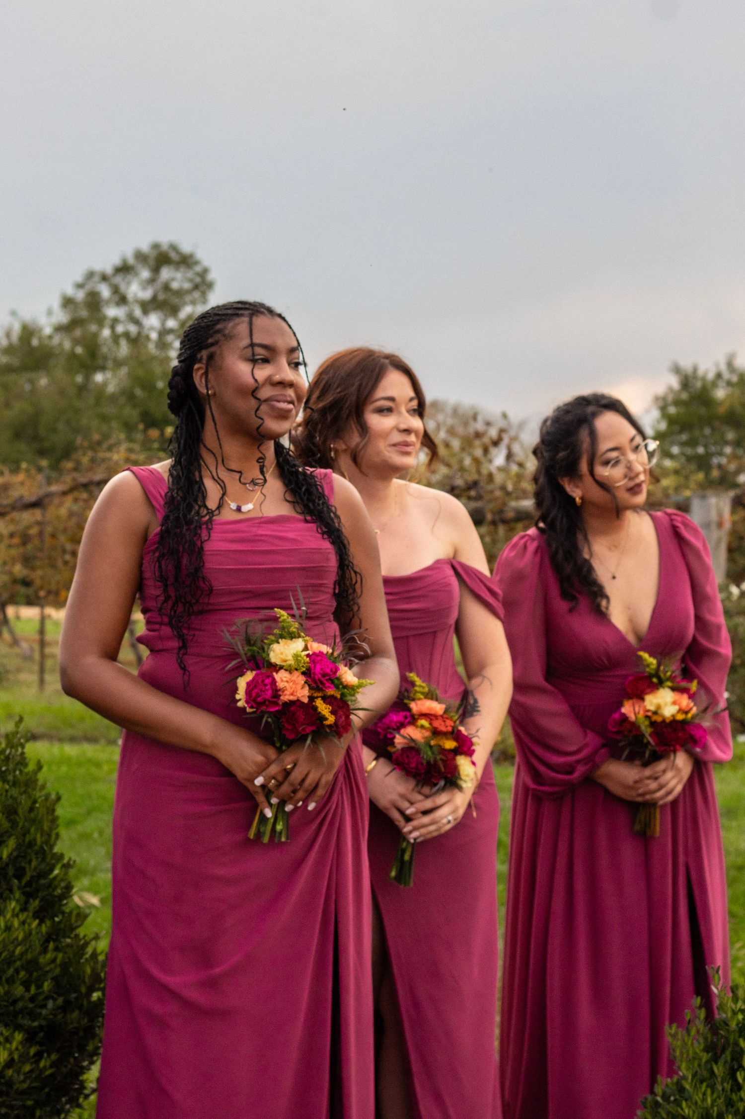 Three bridesmaids in matching magenta gowns holding colorful bouquets stand outdoors at a wedding ceremony.