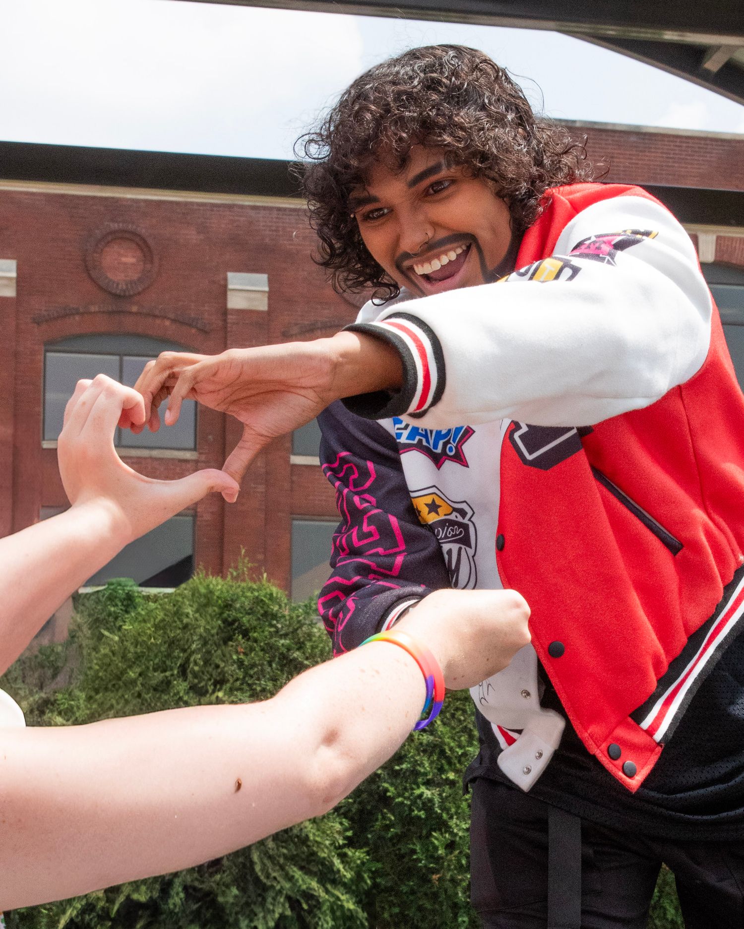 Two people making a heart shape with their hands against a brick building backdrop in a cheerful outdoor setting.