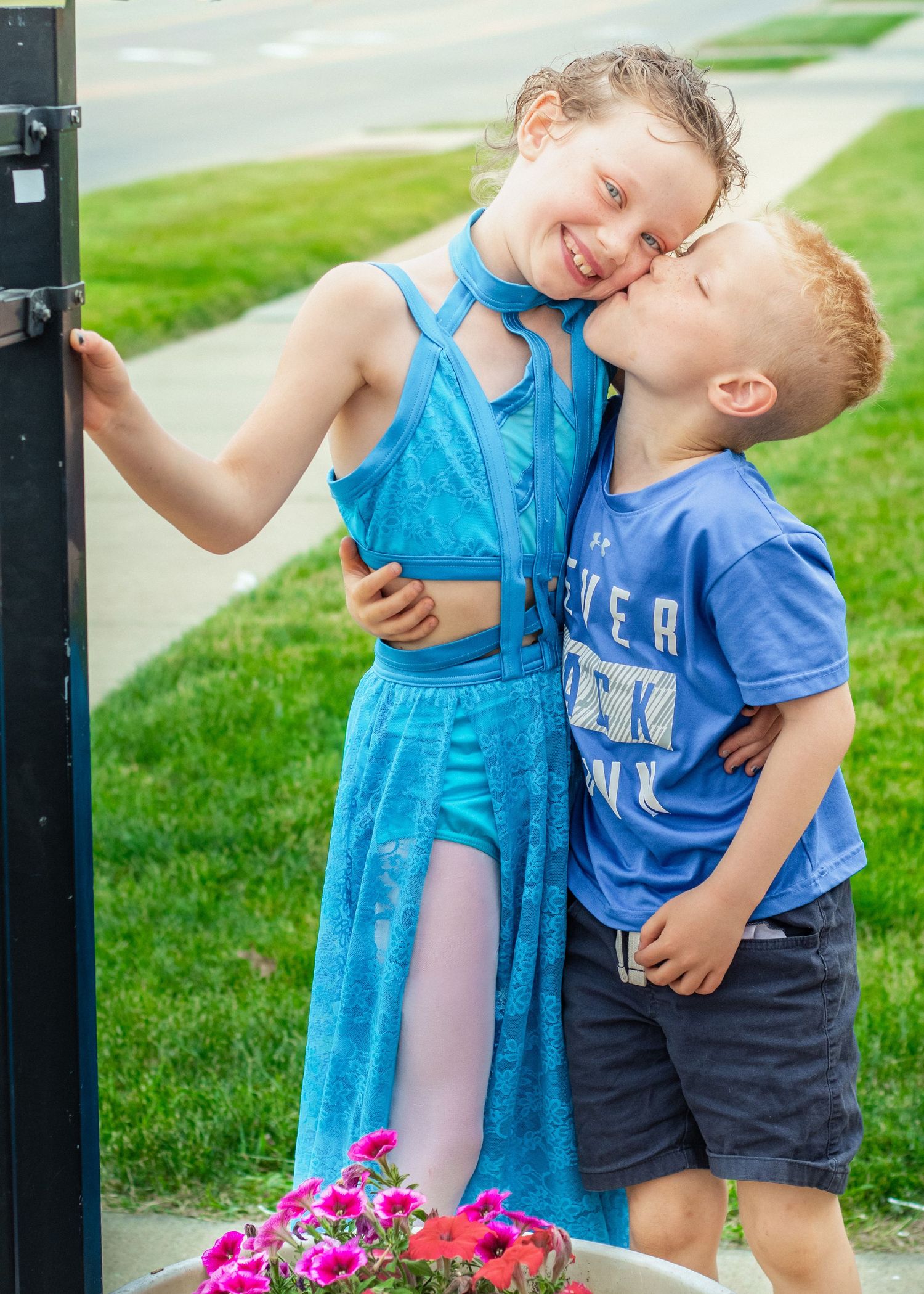 A young boy kisses a smiling girl in a blue dress on the cheek outdoors near pink flowers.