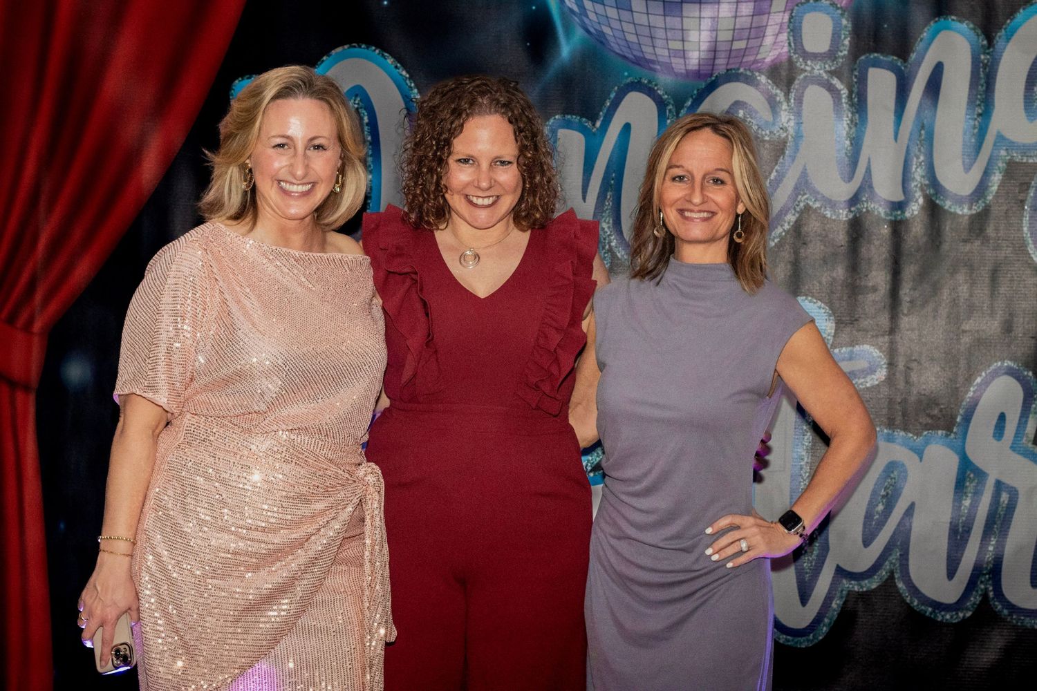 Three women smiling and posing together at a formal gala event with decorative backdrop.