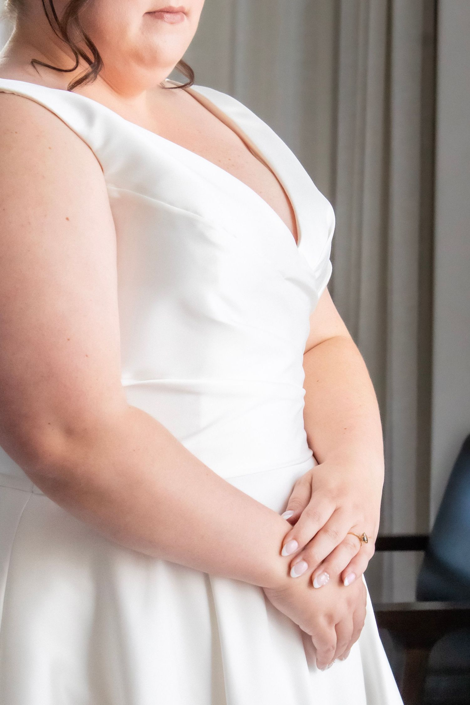 Bride in elegant white V-neck wedding gown clasping hands, showcasing a gold ring, standing gracefully indoors.