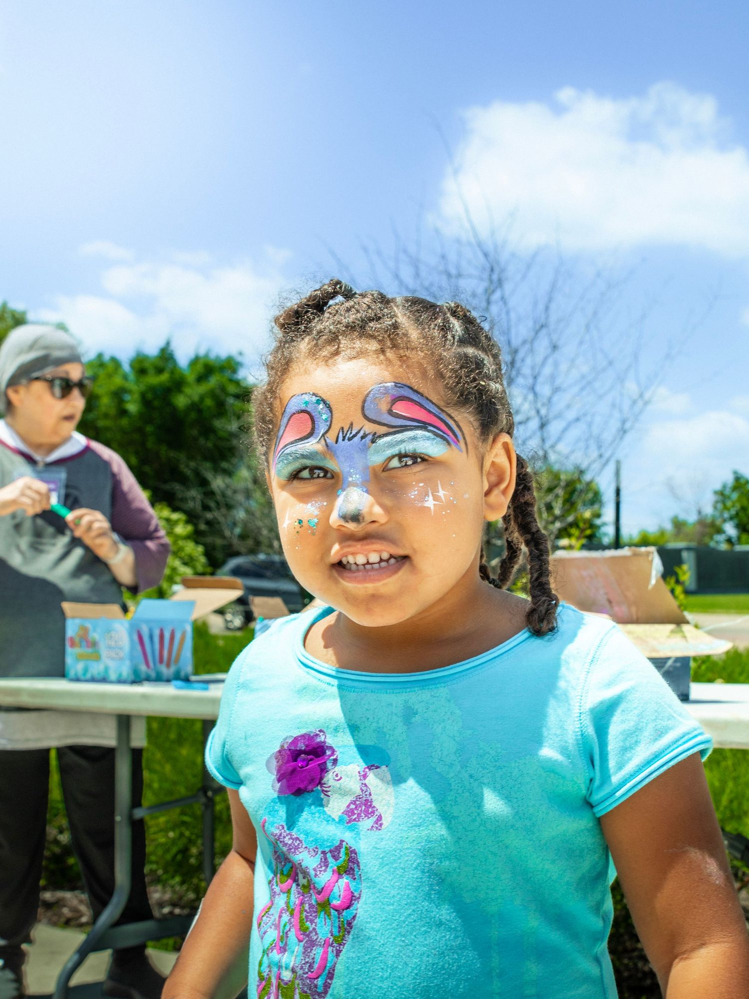 A young girl with colorful butterfly face paint smiles outdoors at a sunny festival event.