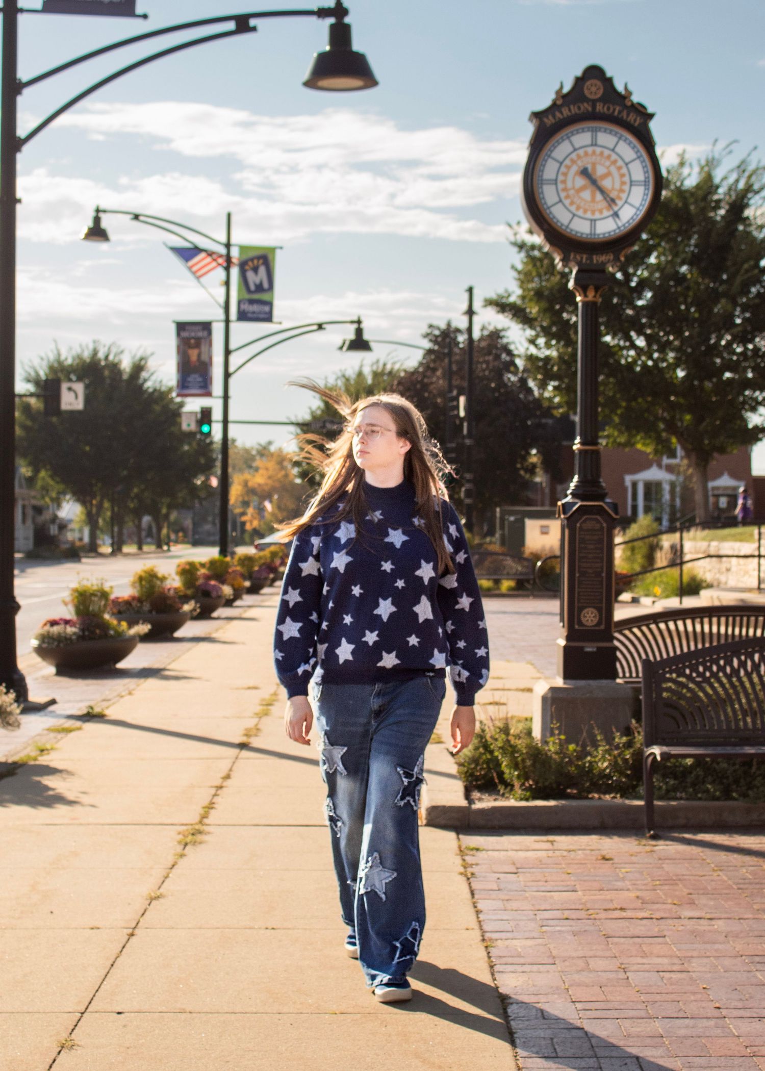 A person in a star-patterned sweater and jeans walks along a brick sidewalk with vintage lampposts and a decorative clock.