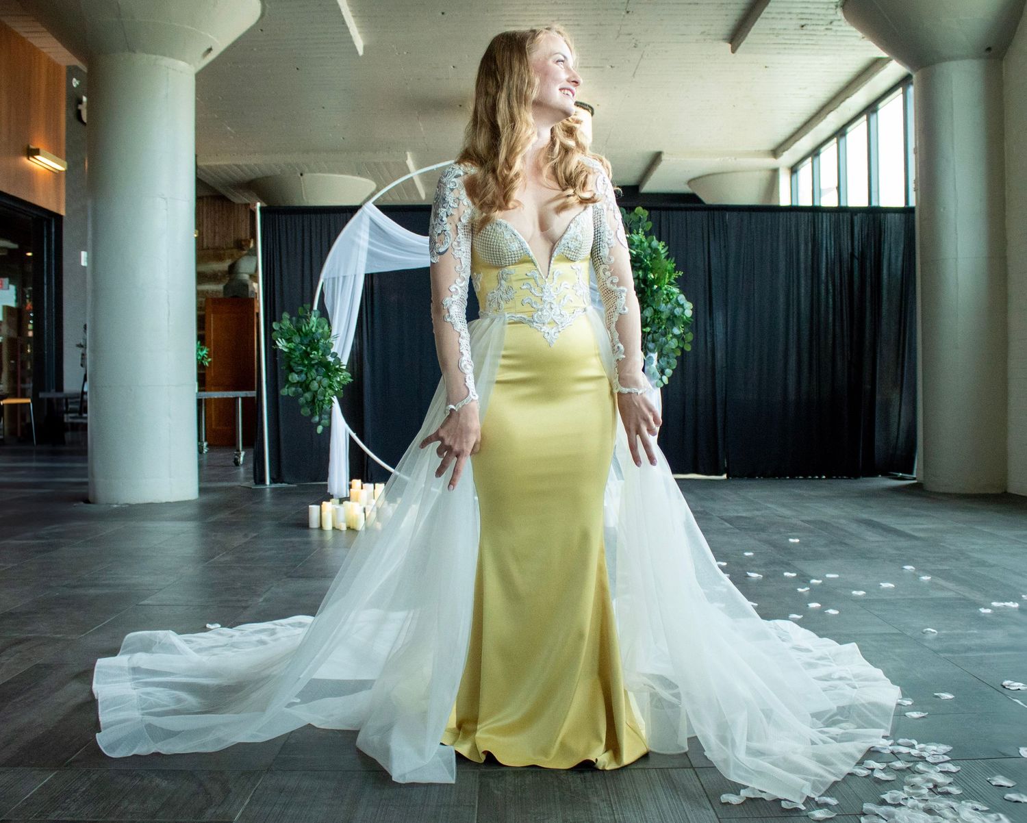 A flowing white wedding dress train trails behind a figure in a yellow dress walking through a modern building hallway.