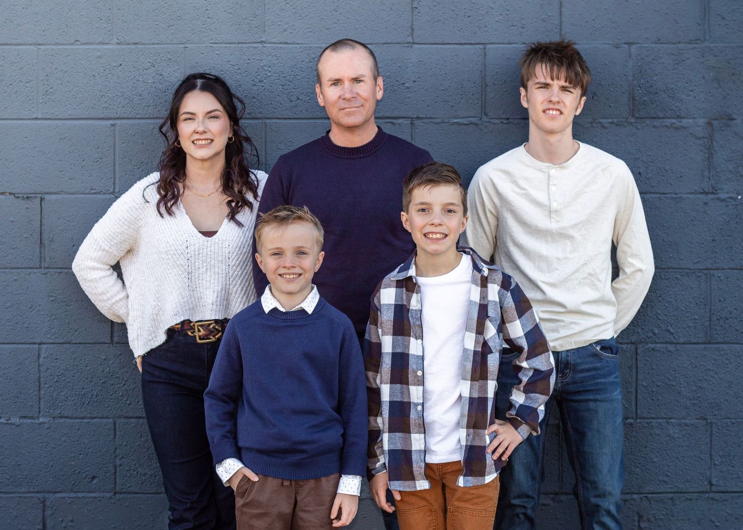 Family group poses against grey concrete wall in coordinated casual outfits.