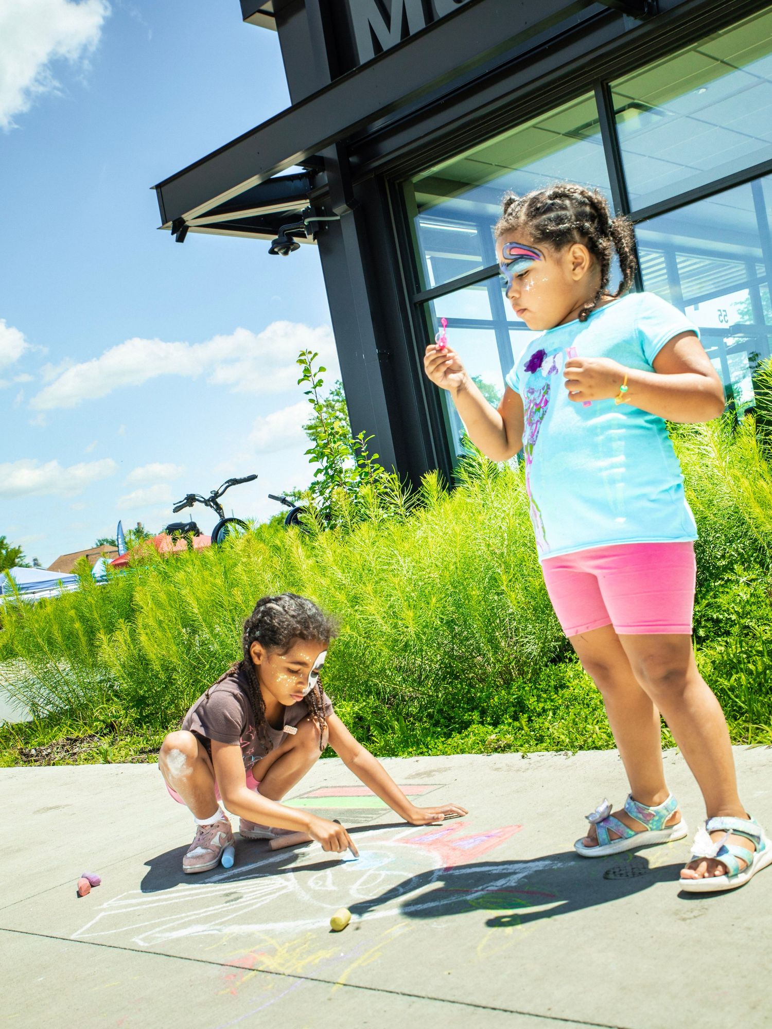 Two young girls drawing colorful chalk art on a sidewalk outside a building on a sunny summer day.