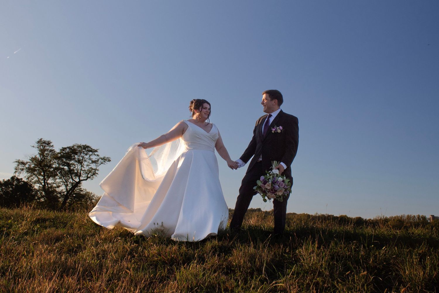 Bride in white gown and groom in suit hold hands walking through green field under clear blue sky.