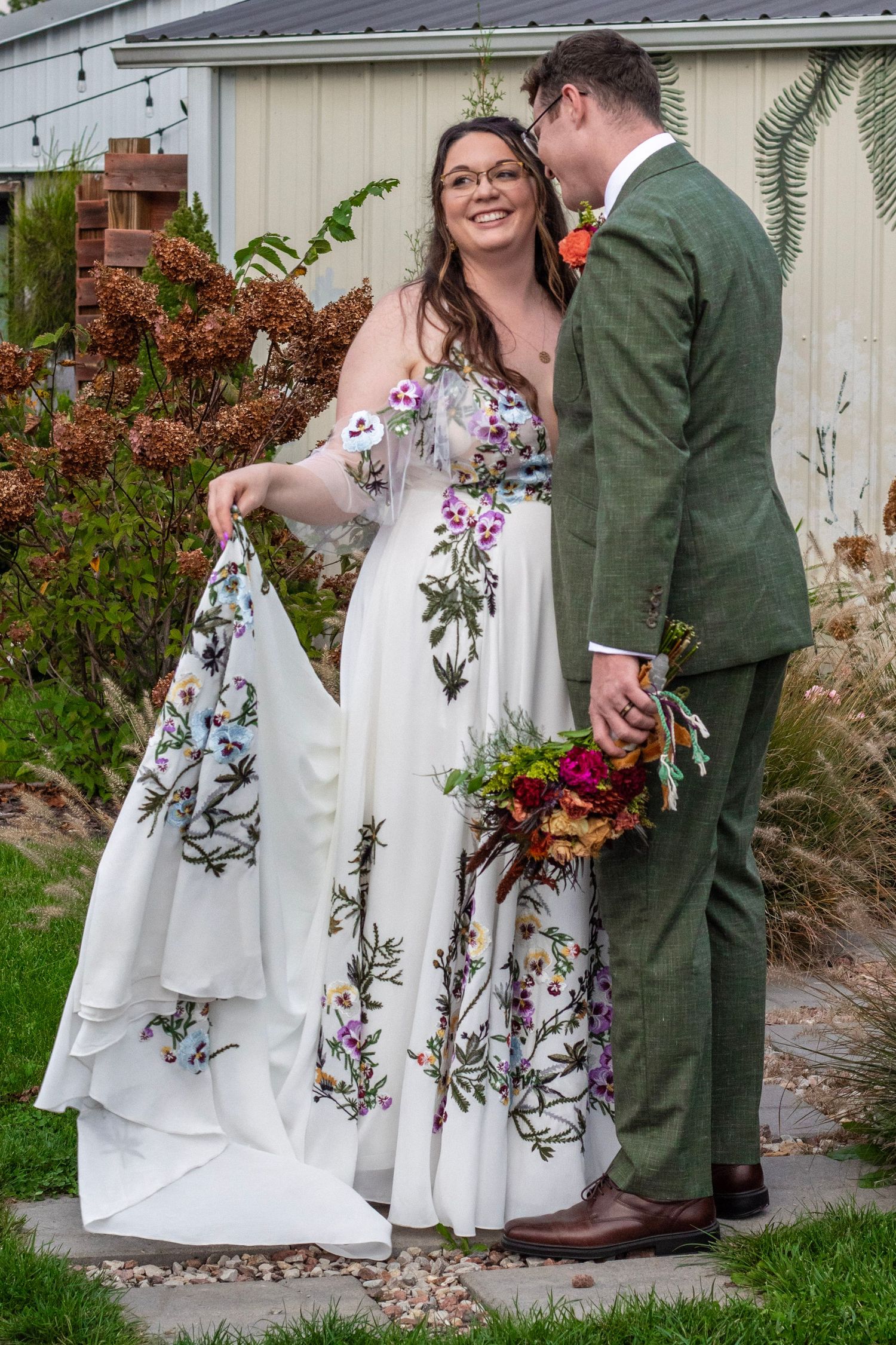 Bride in floral wedding gown holding train stands with groom in green suit holding bouquet outdoors.