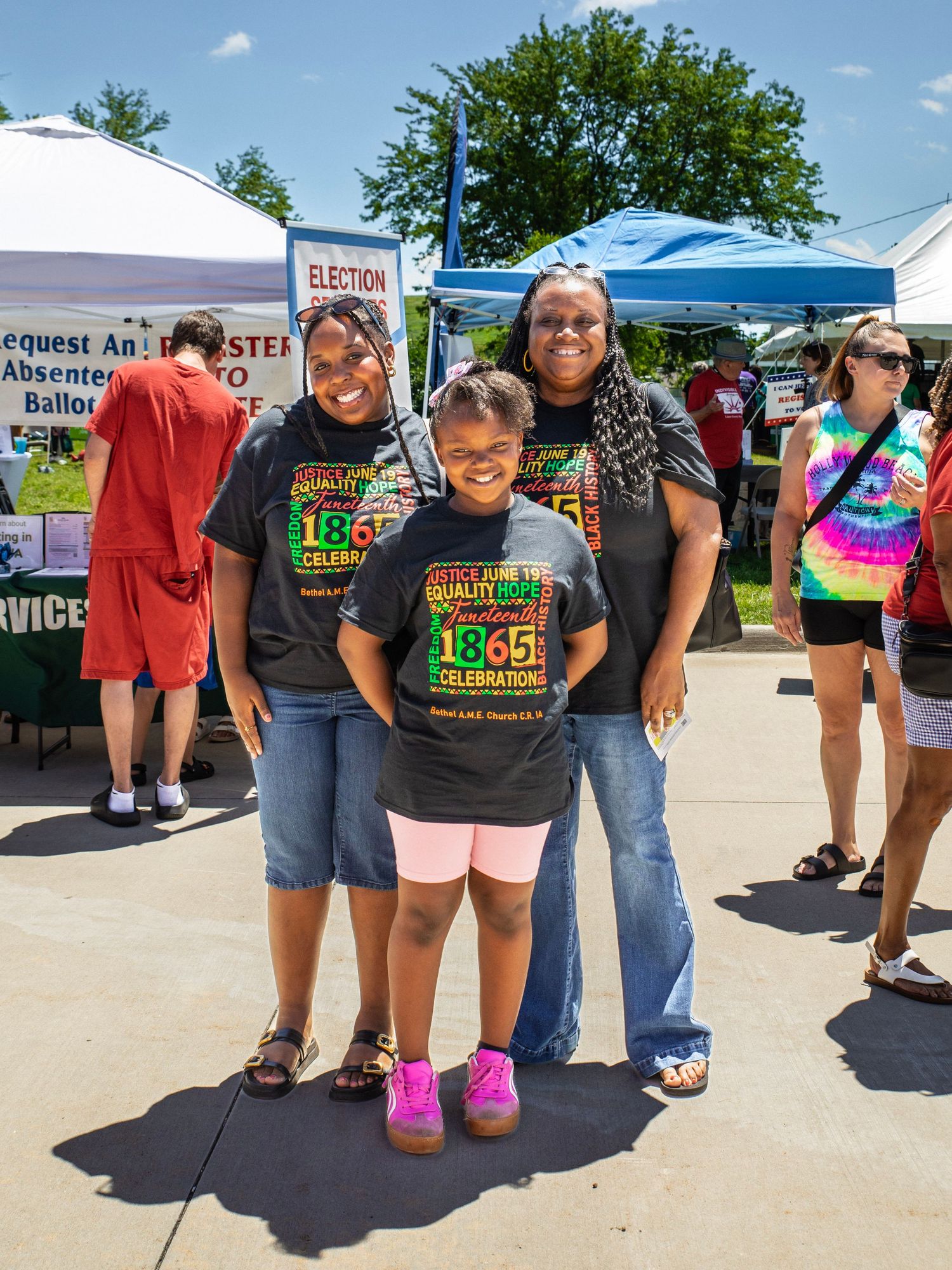 Three people wearing Juneteenth 1865 celebration t-shirts pose together at an outdoor community event on a sunny day.