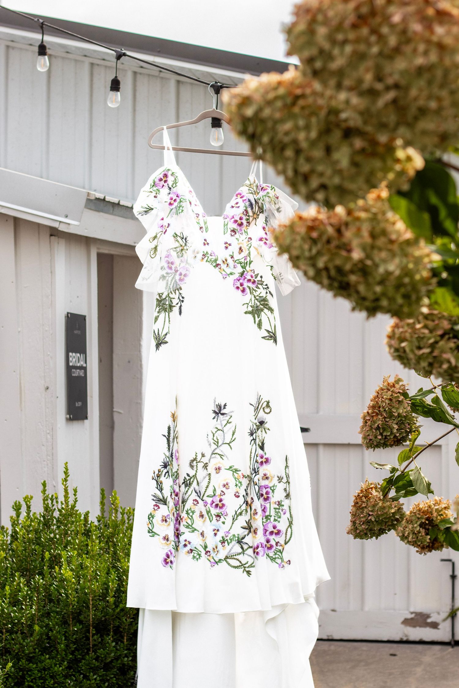 White floral embroidered wedding dress hanging outdoors near a bridal boutique with hydrangeas.