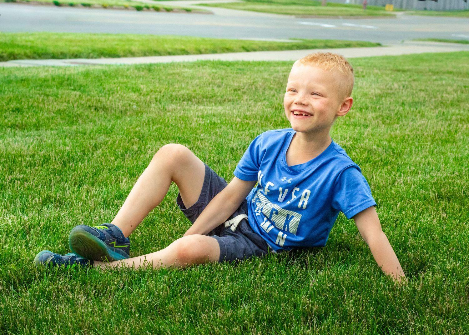Smiling young blonde boy sitting on lush green grass wearing a blue t-shirt and shorts on a sunny day.