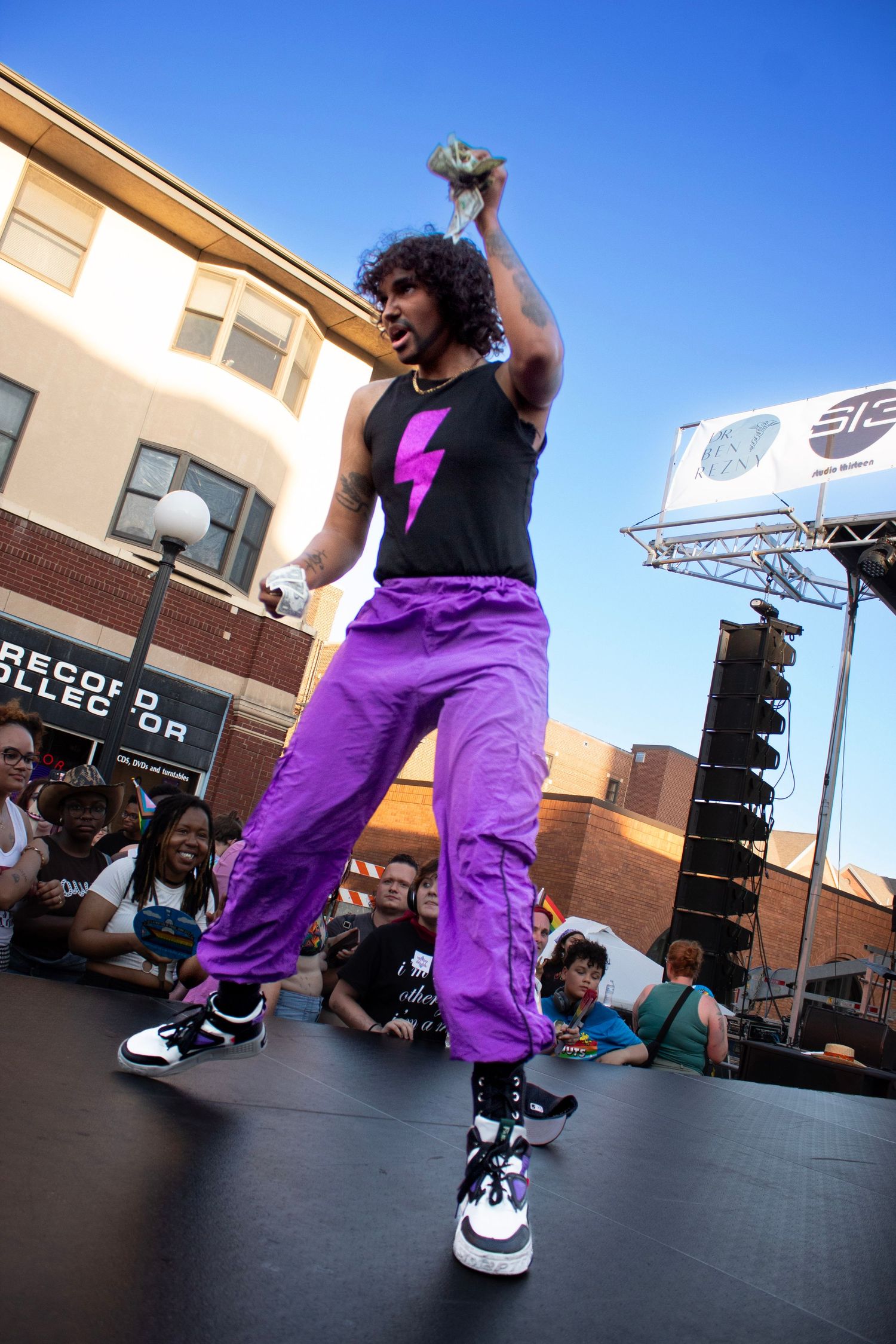 Energetic performer in black tank top and purple pants dancing on outdoor stage.