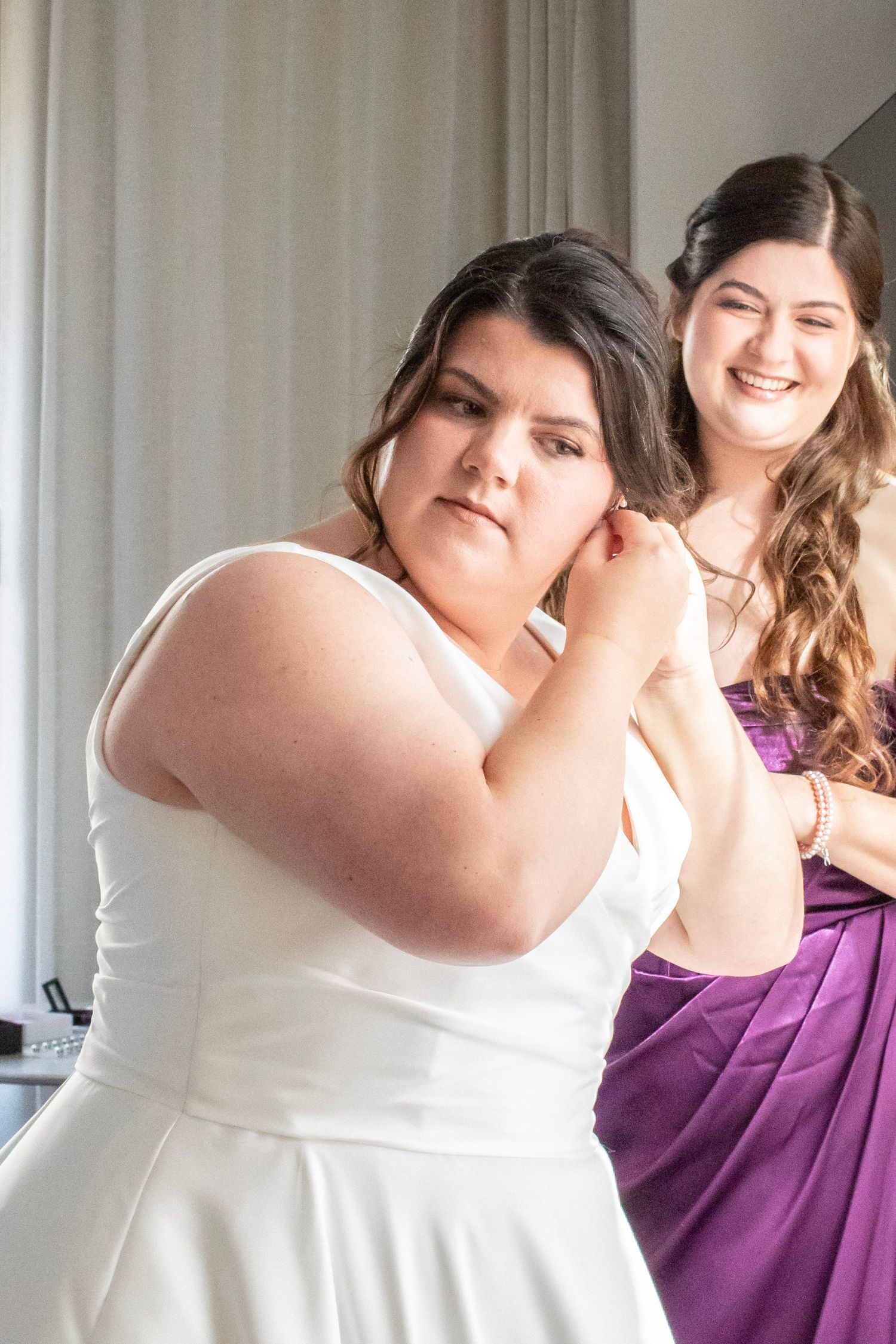 Bride in white dress putting on earring assisted by smiling bridesmaid in purple dress during wedding preparations.