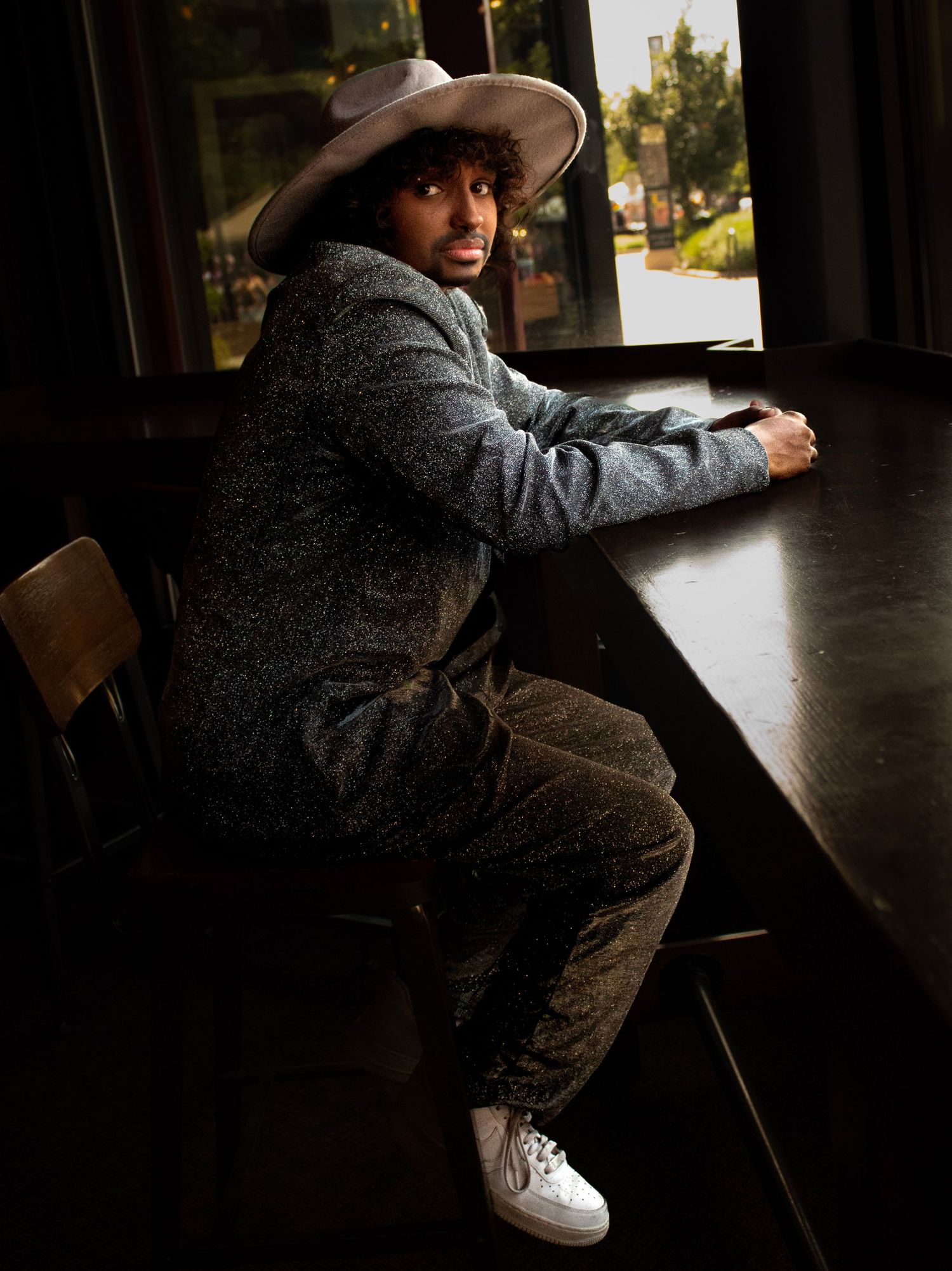 Person in a cowboy hat and grey sweater sits on a wooden chair in a dark interior space with natural lighting.