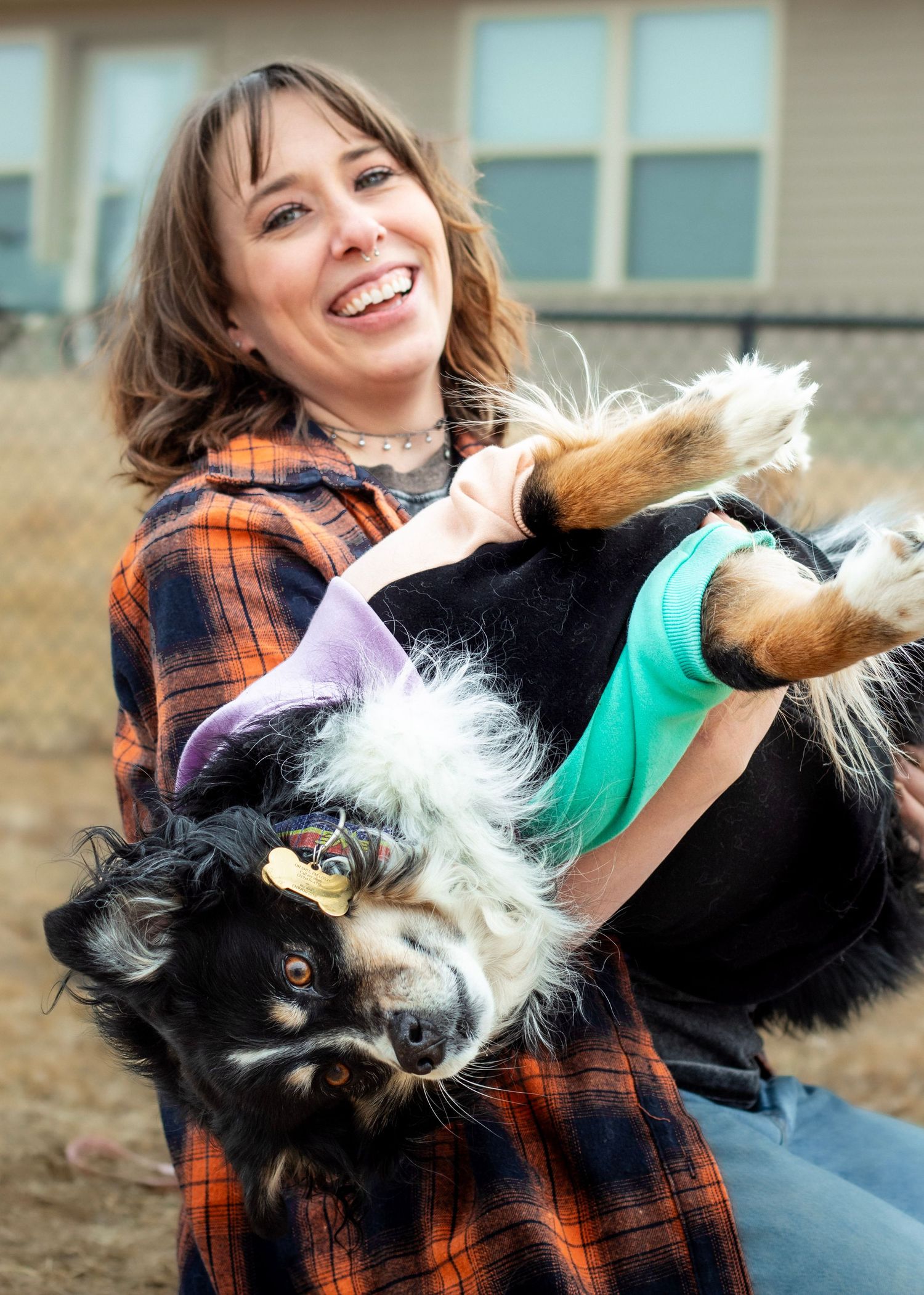 Smiling woman in plaid shirt playfully holding a black and white dog outdoors near a building.