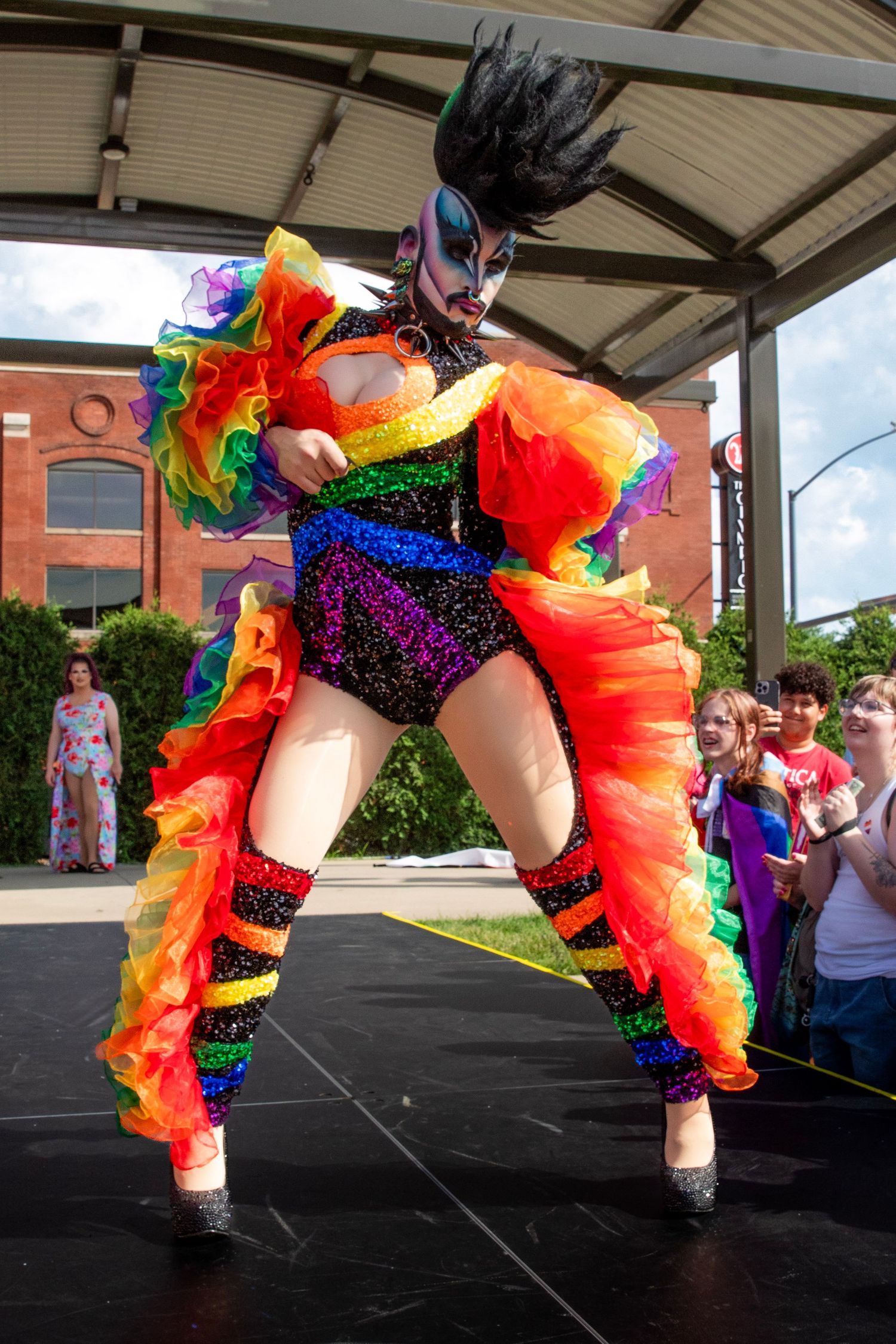 Dancer in colorful rainbow tutu and sparkly costume performs energetic splits during outdoor performance.