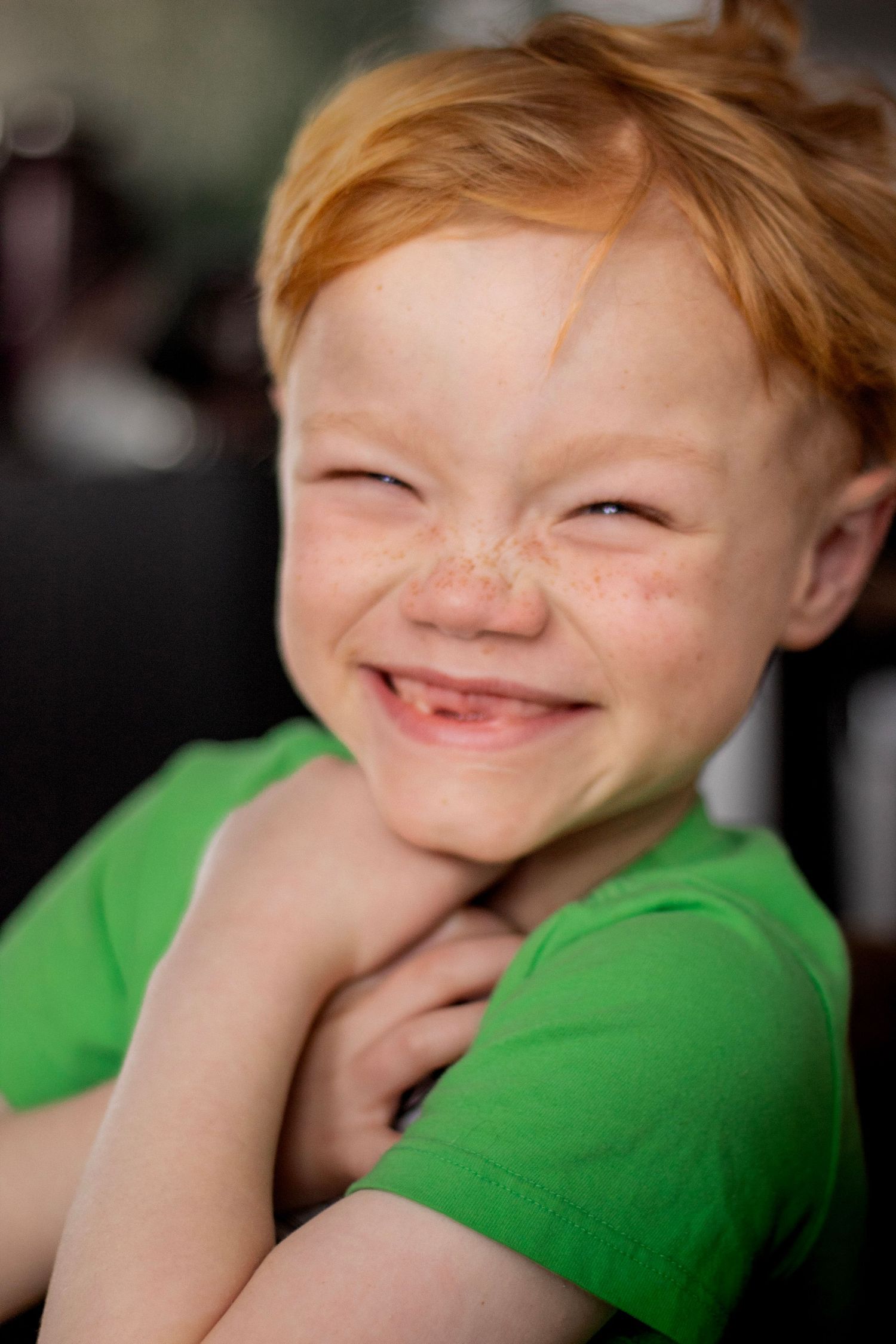 A cheerful red-haired freckled toddler in a green shirt squinting and smiling broadly with joy.