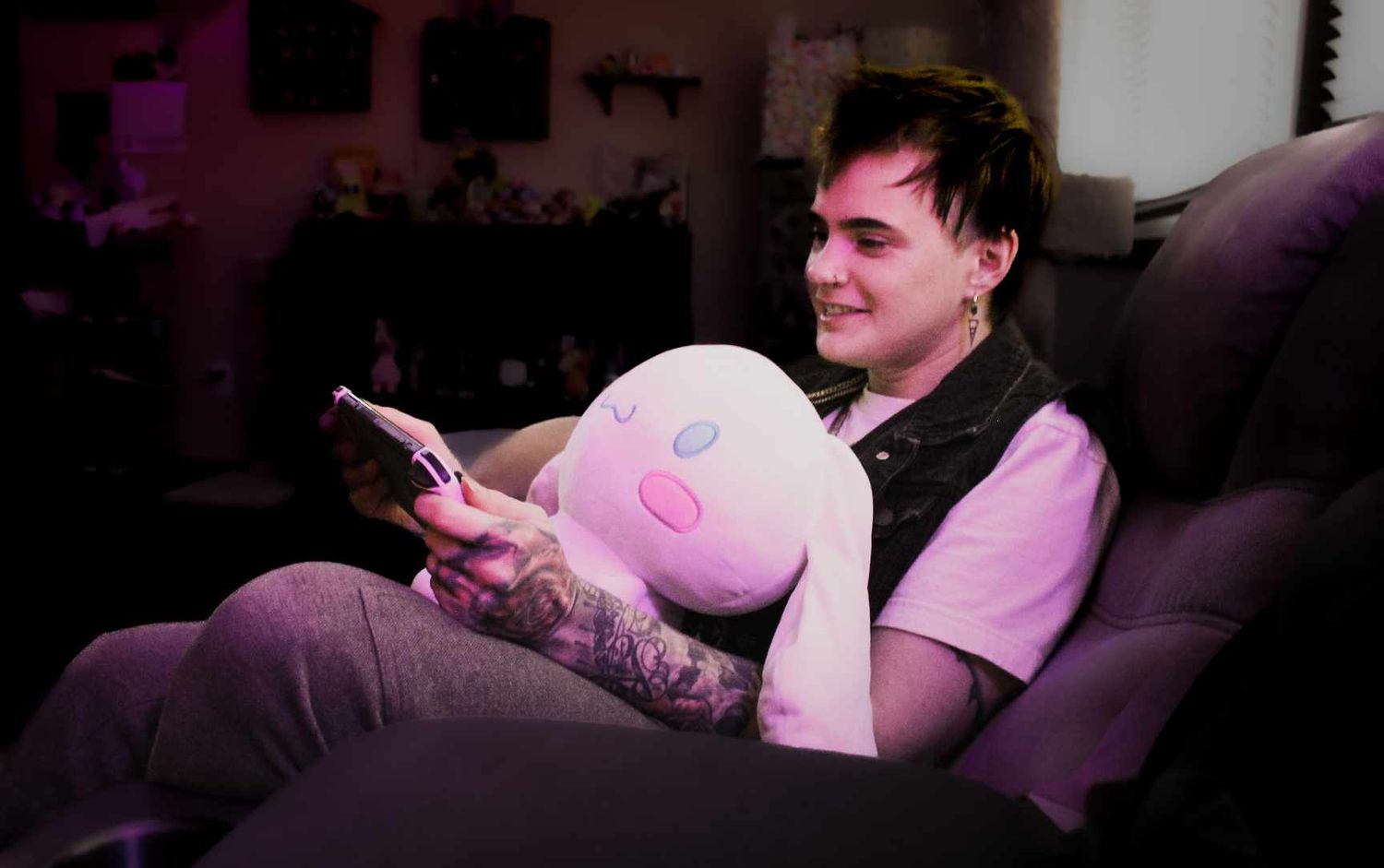 Person relaxing on couch with phone and pink plush toy in dimly lit room.
