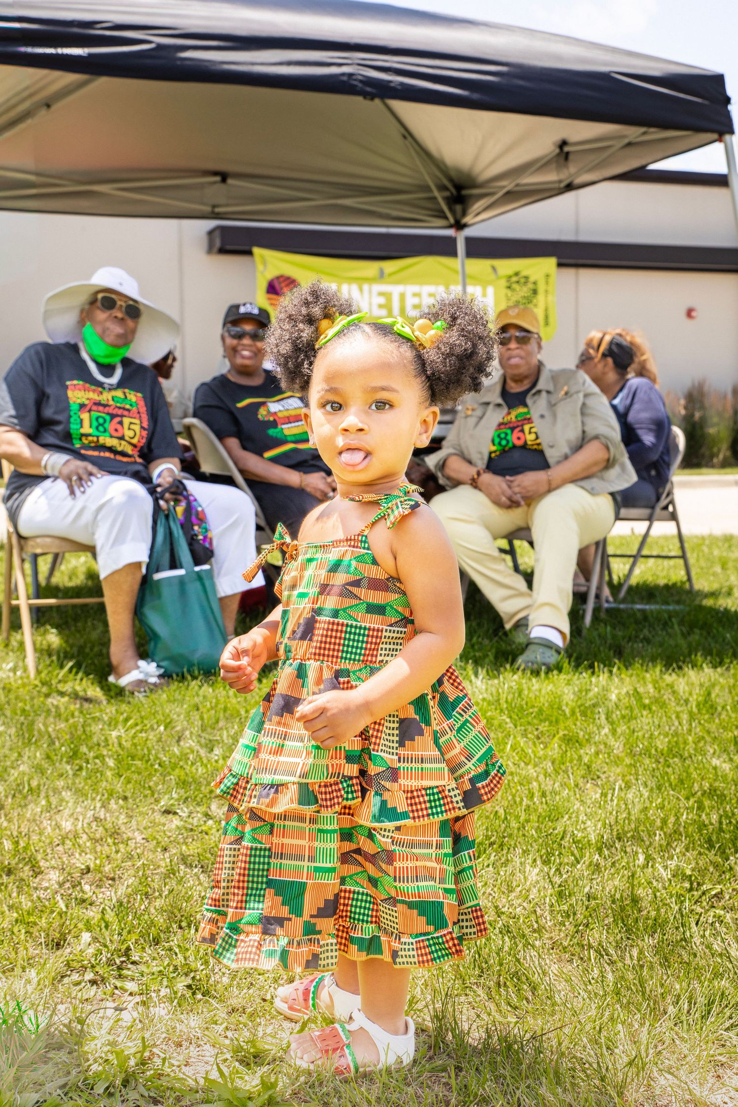 Young girl in colorful kente dress dancing at a Juneteenth celebration outdoors with family seated under a canopy.