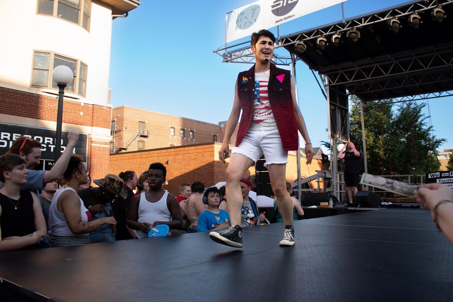 Performer in maroon vest and shorts on outdoor stage with audience seated.