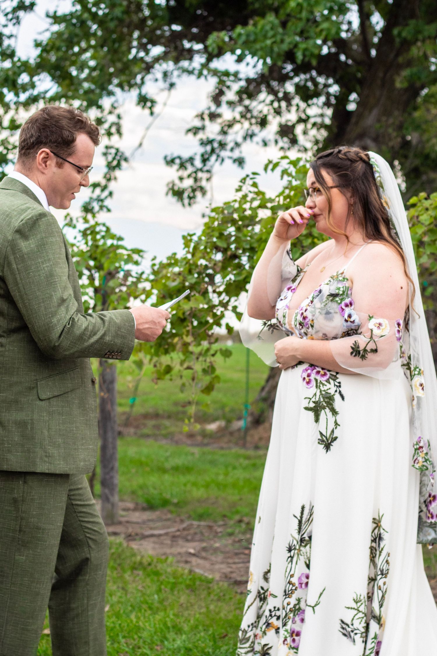 Emotional bride in floral wedding dress wipes tear as groom reads vows outdoors in lush green garden setting.