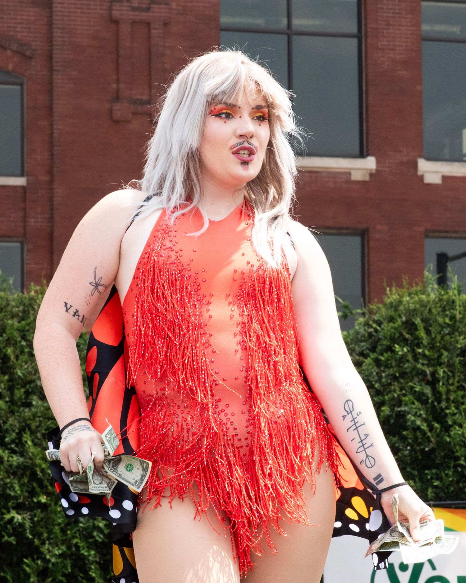 A person in a bright coral fringe bodysuit poses energetically against a brick building exterior.
