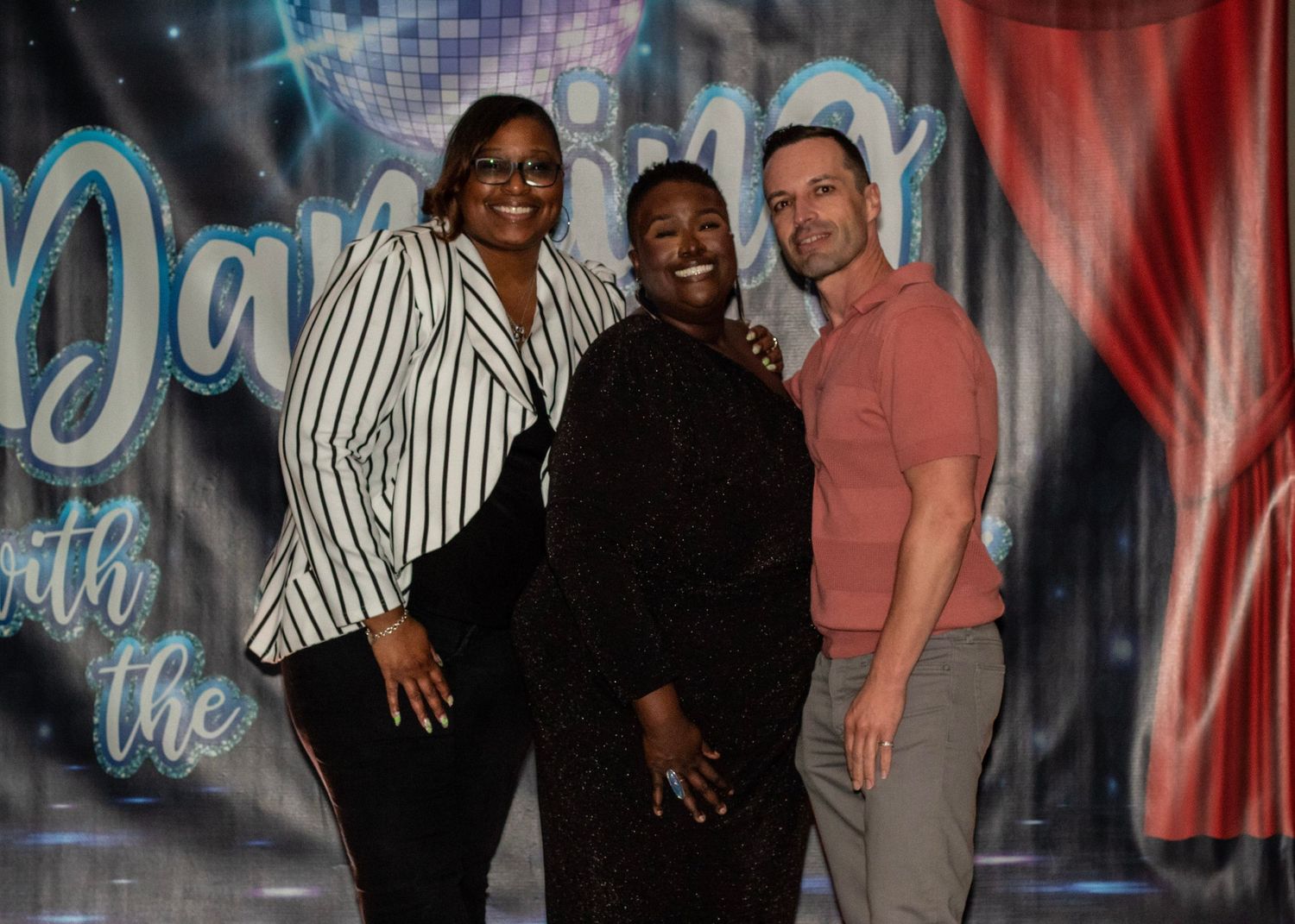 Three people smiling together at a Dancing with the Stars themed event backdrop.